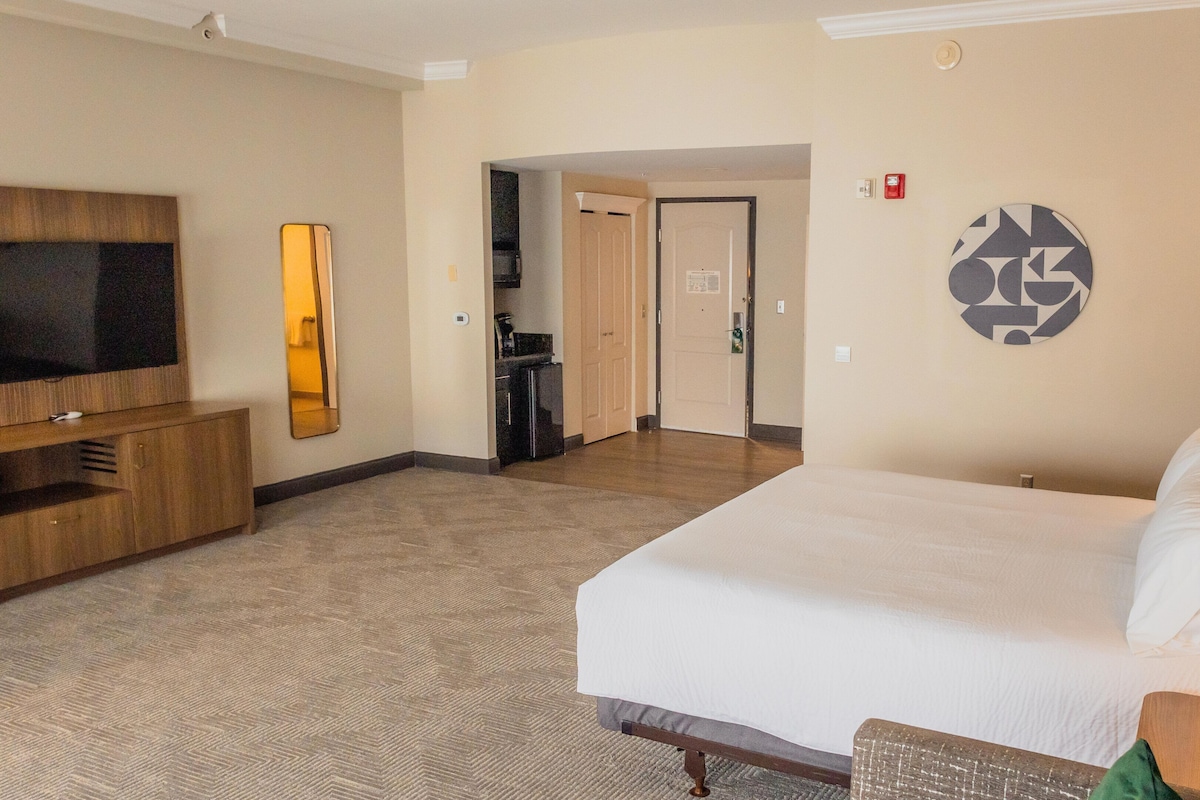 A hotel room with a large bed, a TV, a mirror, a kitchenette, and a door leading to another room.