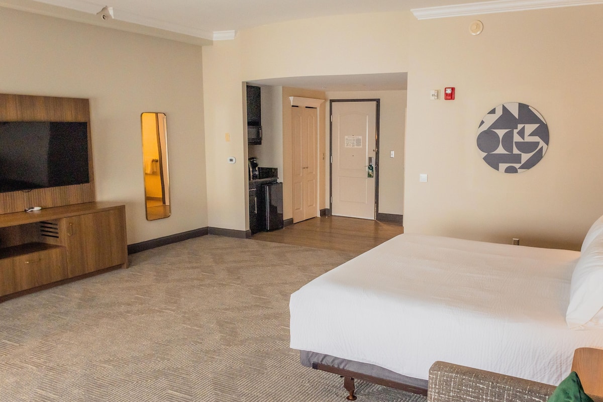 A hotel room with a large bed, a TV, a mirror, a kitchenette, and a door leading to another room.