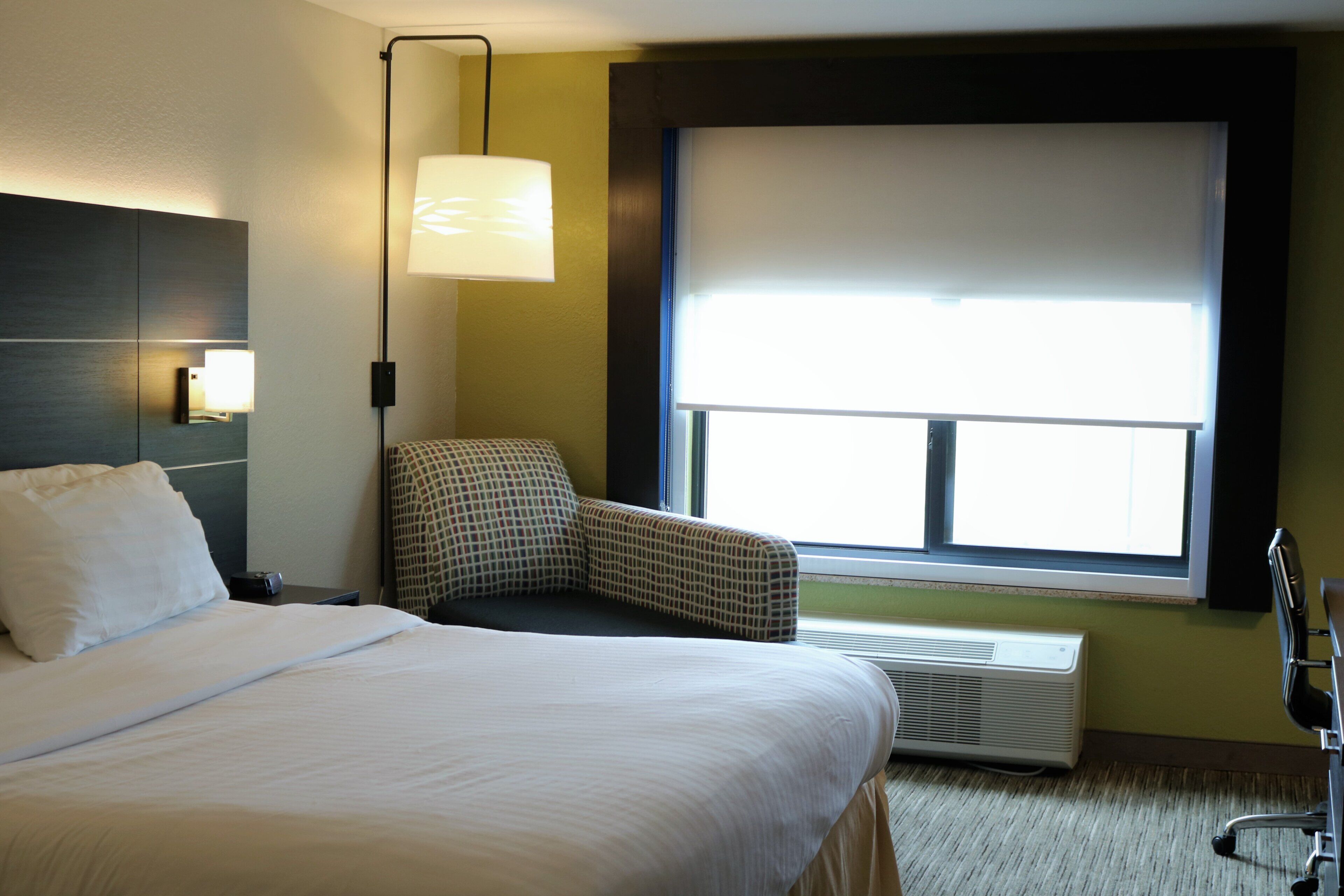 A hotel room with a bed, a chair, a window with blinds, a wall-mounted lamp, and a radiator.