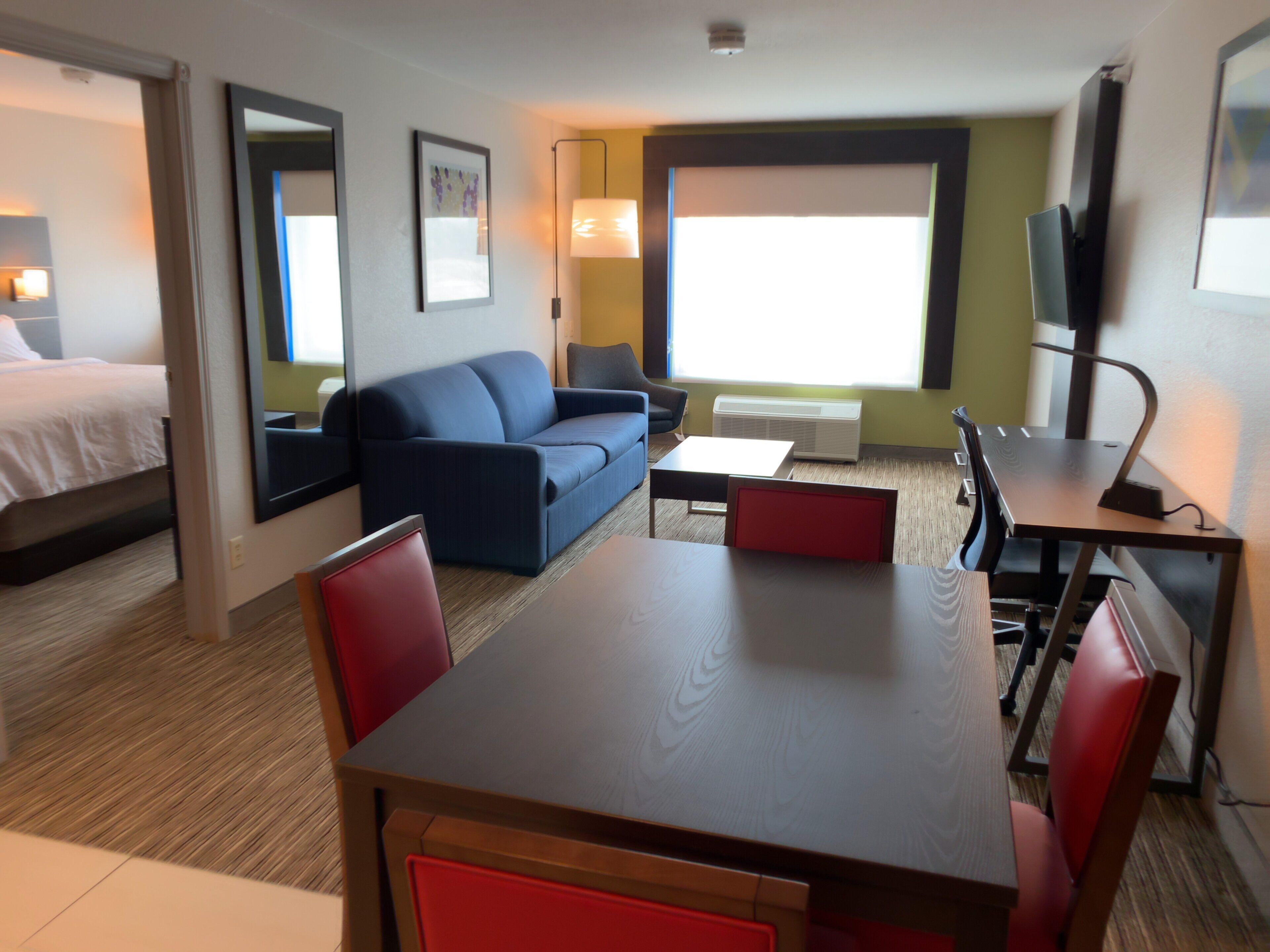 Habitación de hotel con cama, sofá, mesa de comedor con sillas, televisor y ventana con persianas.