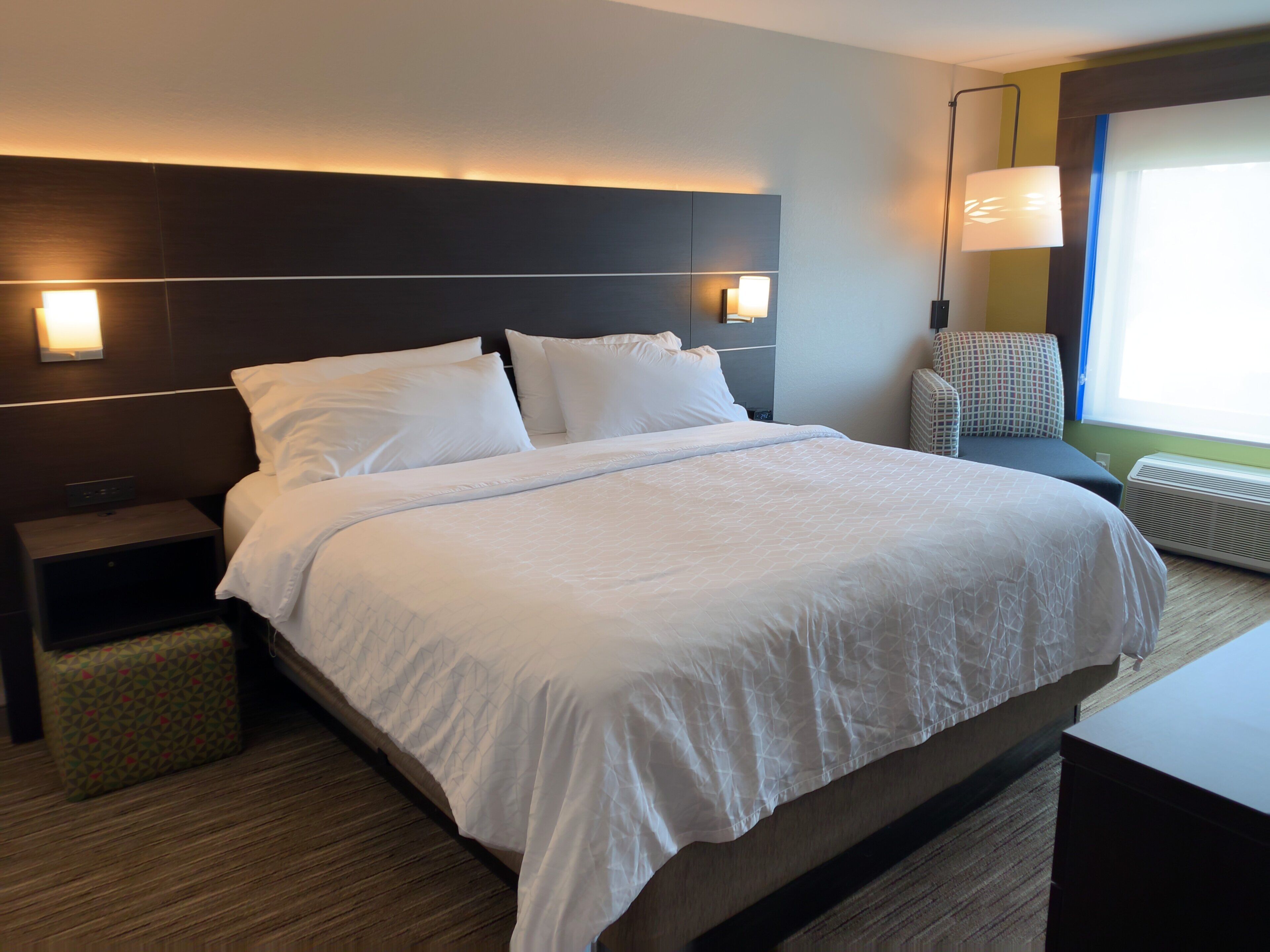 Una habitación de hotel con una cama grande, una mesita de noche, una lámpara, una silla y una ventana.