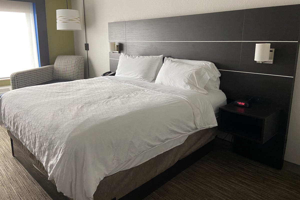 A hotel room with a large bed, a nightstand with a clock, a chair, and a lamp.