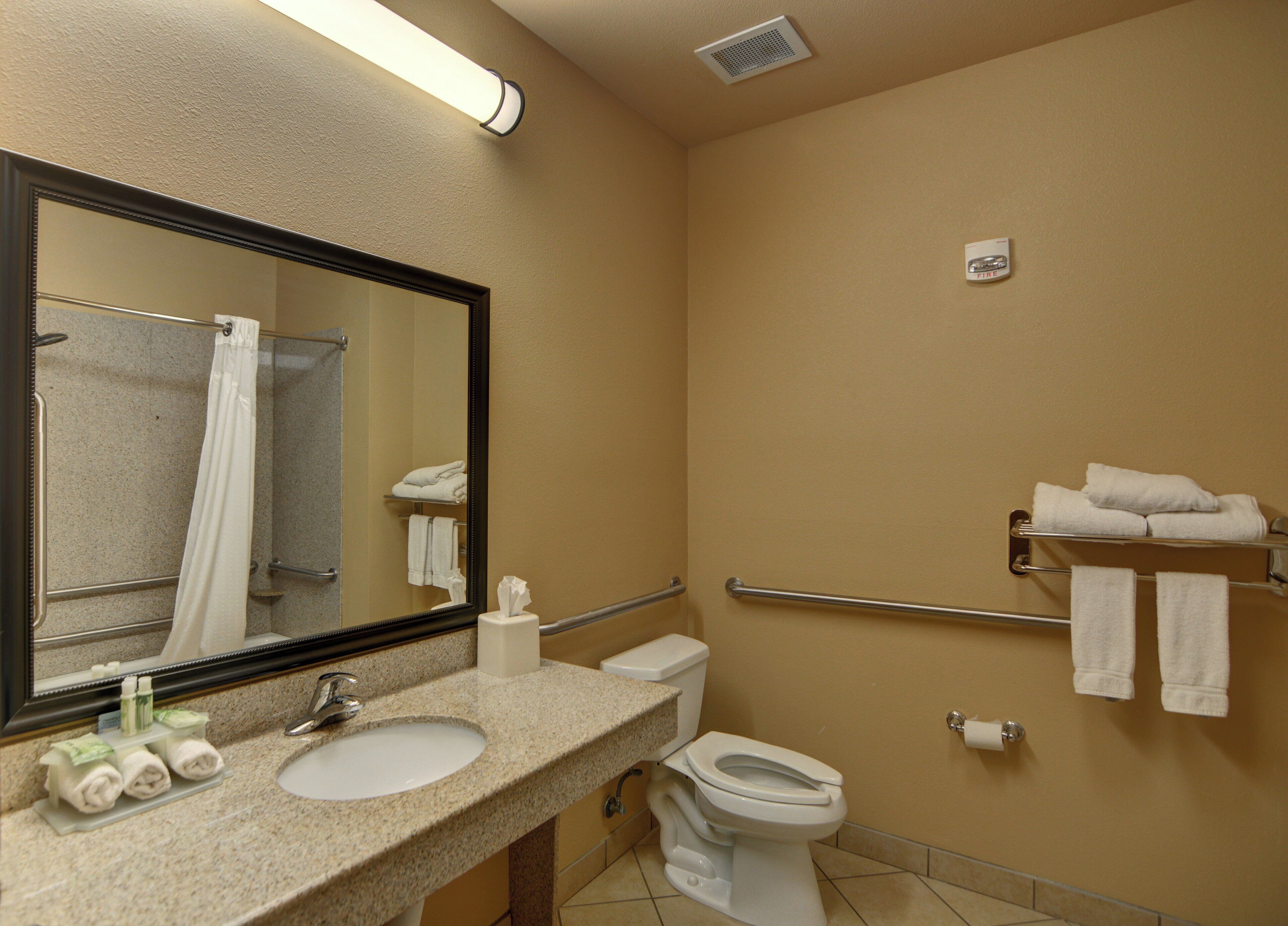 A bathroom with a sink, toilet, and a mirror. There are towels on a rack and a fire extinguisher on the wall.