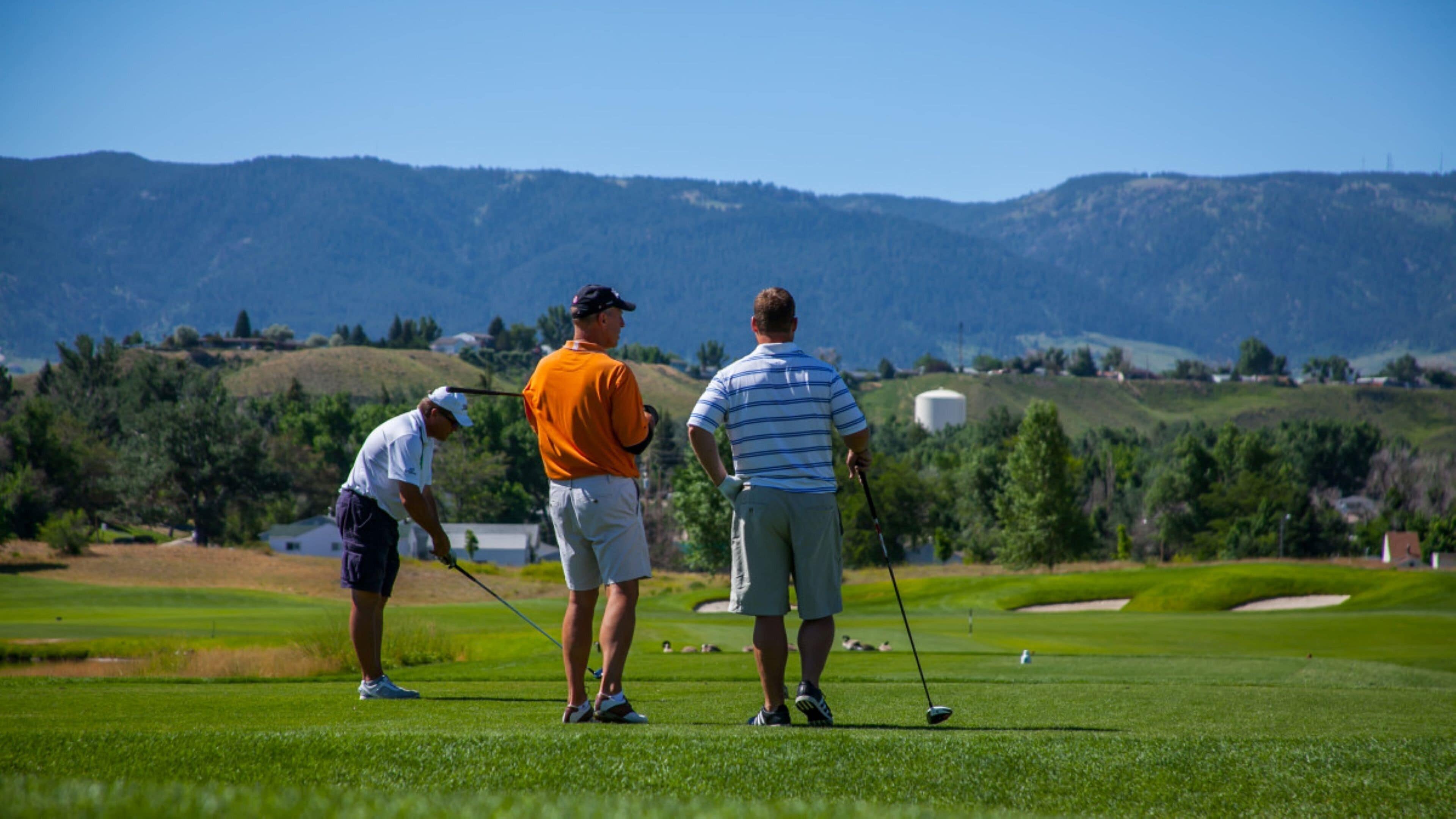 Tres personas en un campo de golf, con montañas al fondo.
