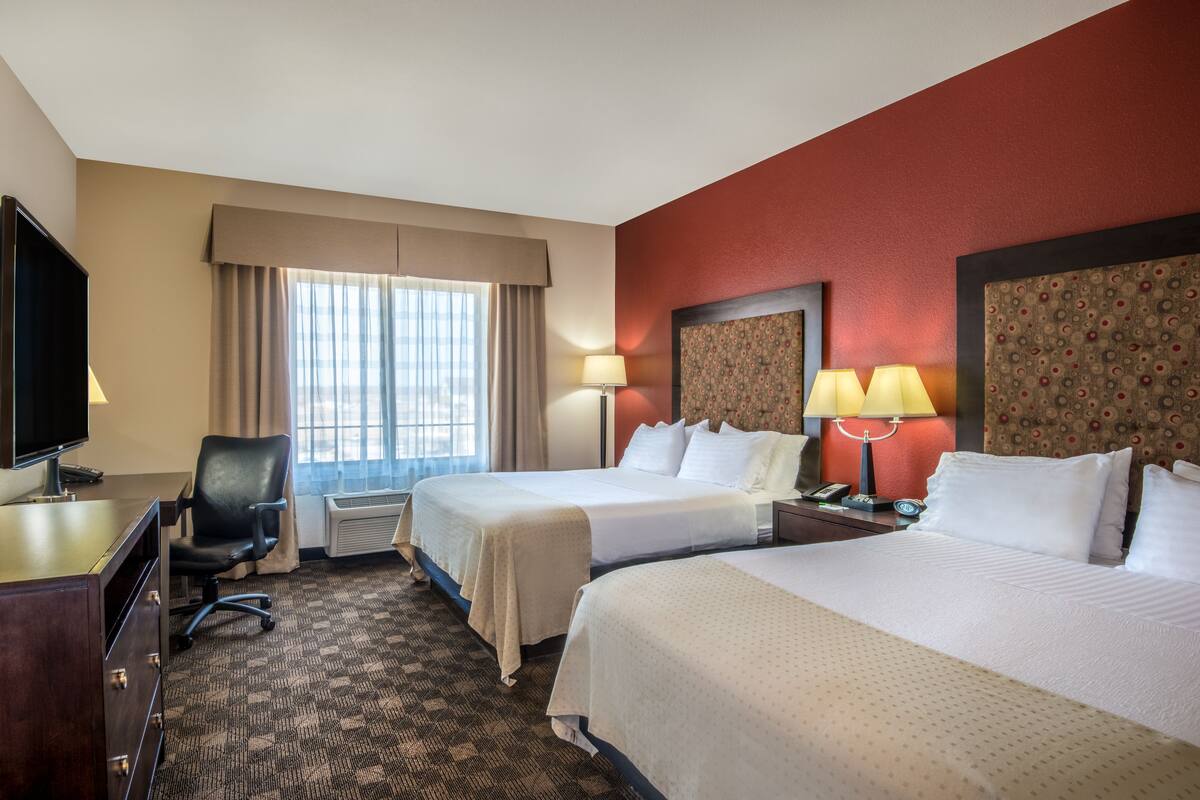 Quarto de hotel com duas camas, uma escrivaninha com televisão, uma cadeira e uma janela com cortinas.