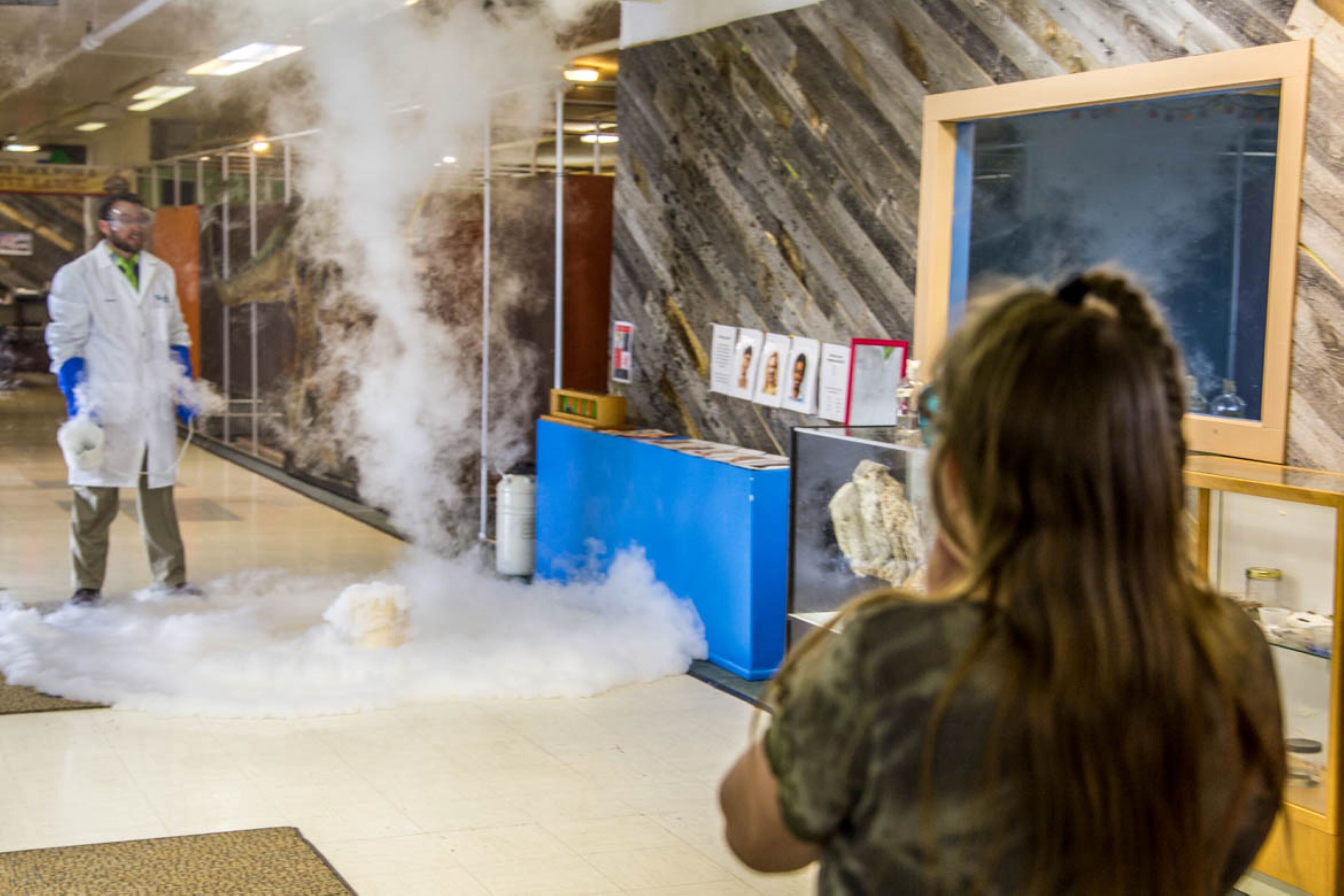 A person in a white lab coat standing next to a blue counter with a display case, surrounded by smoke.