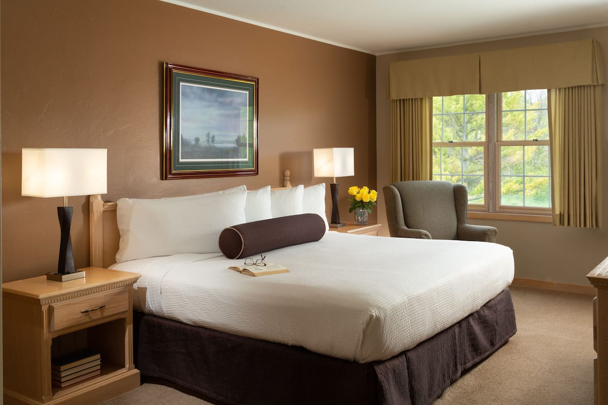 A hotel room with a large bed, two bedside tables, a chair, a window with curtains, and a framed picture on the wall.