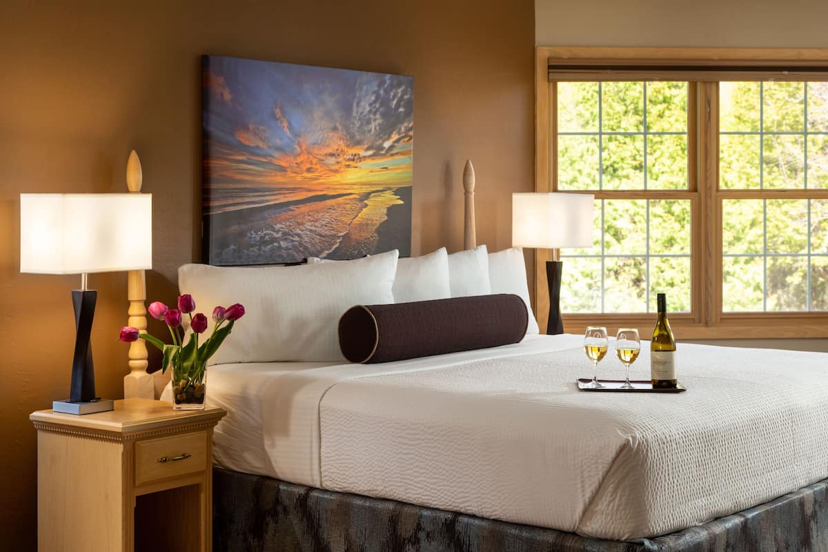 A hotel room with a large bed, two bedside lamps, a painting of a sunset, a vase with flowers, and a tray with wine glasses and a bottle.