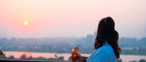 A person holding a drink on a balcony with a view of a sunset over a body of water.