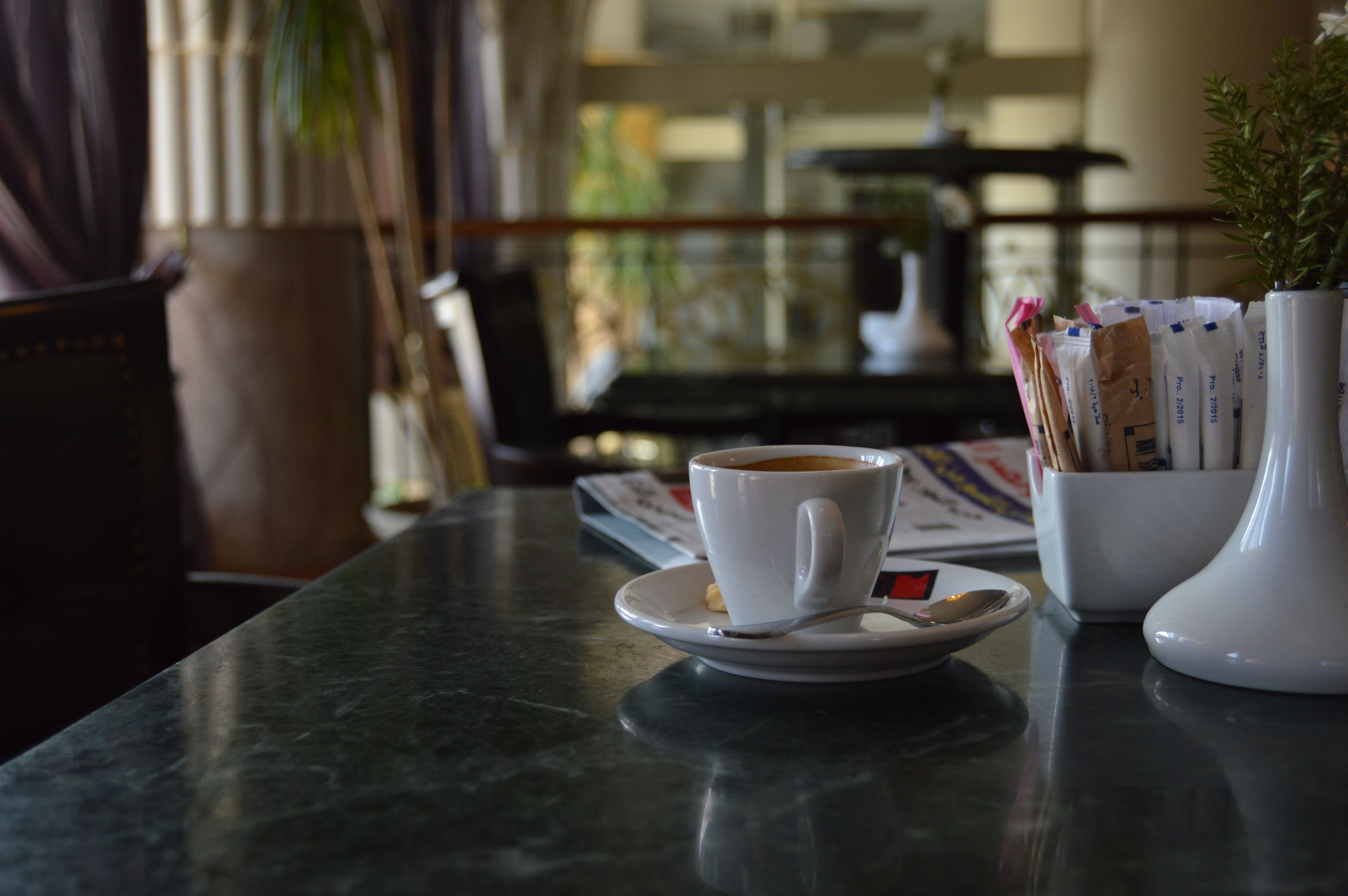 Una mesa con una taza de café, una cuchara, un platillo y un jarrón blanco con una planta.
