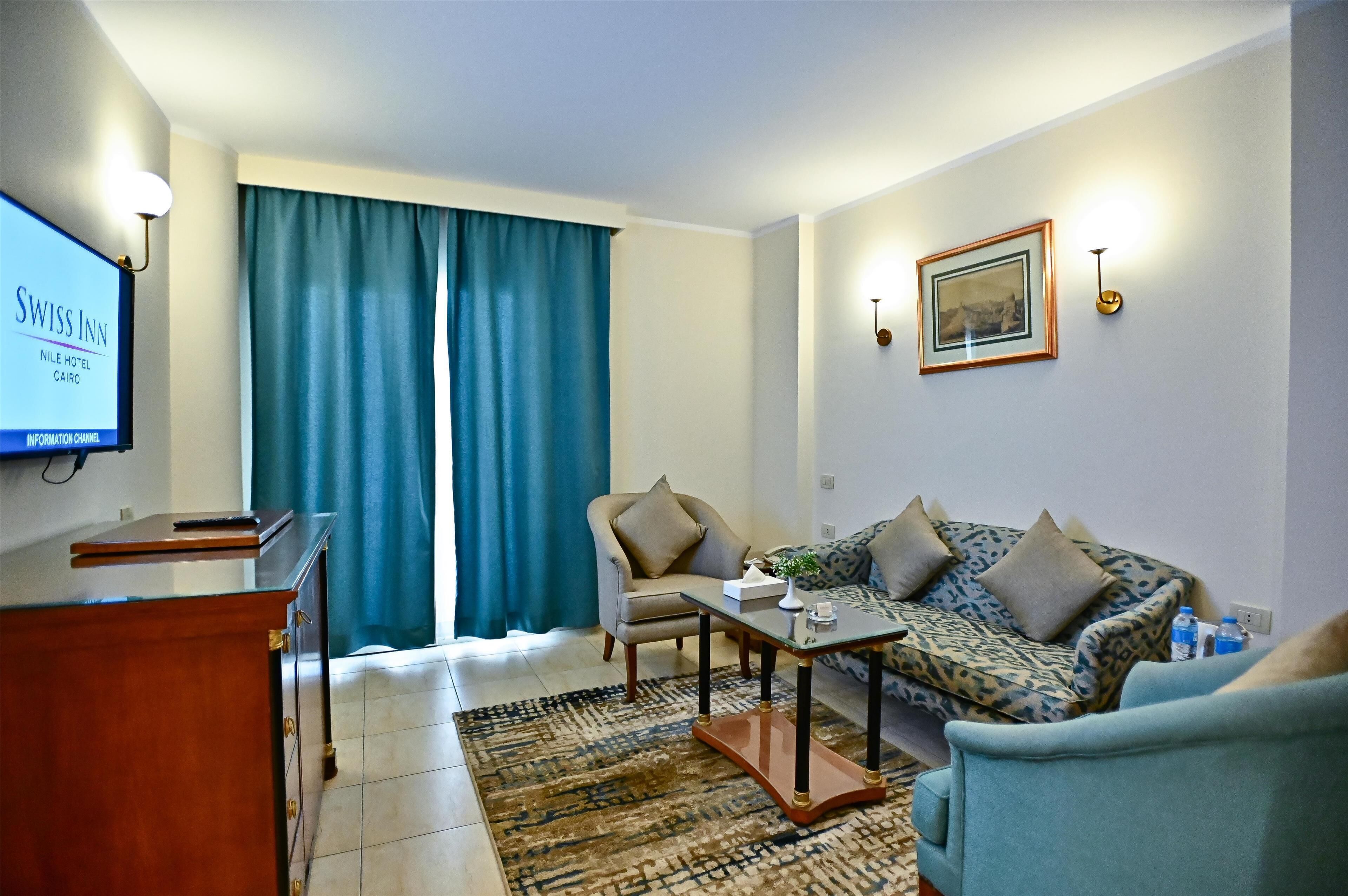 Una habitación de hotel con escritorio, sofá, alfombra estampada y un ventanal con cortinas.
