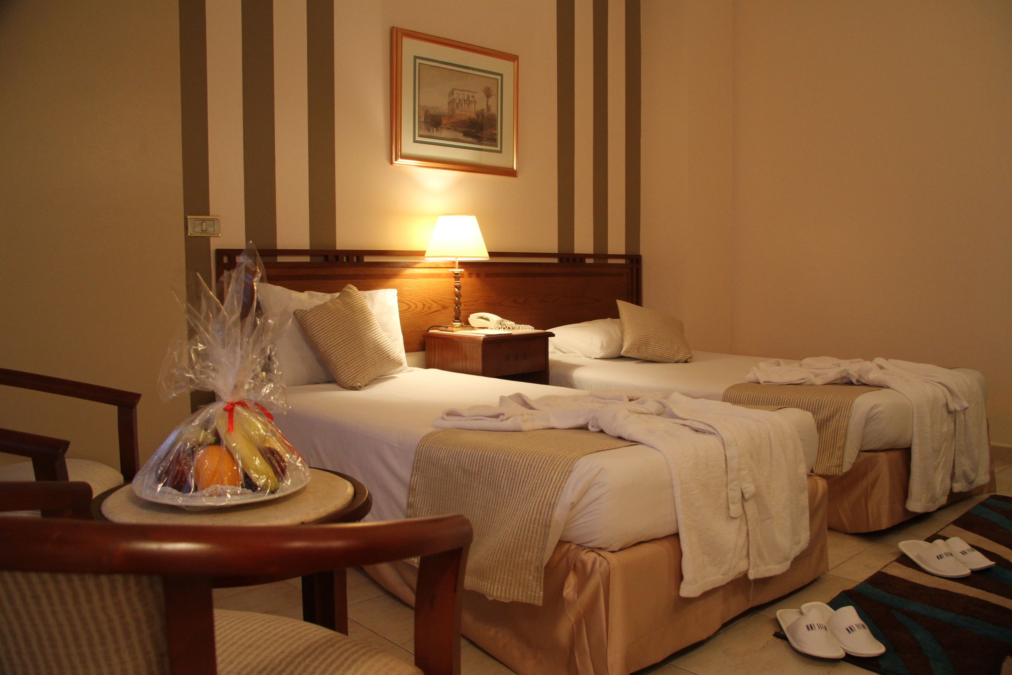 A hotel room with two beds, a bedside table with a lamp, a framed picture on the wall, and a small table with a fruit basket.