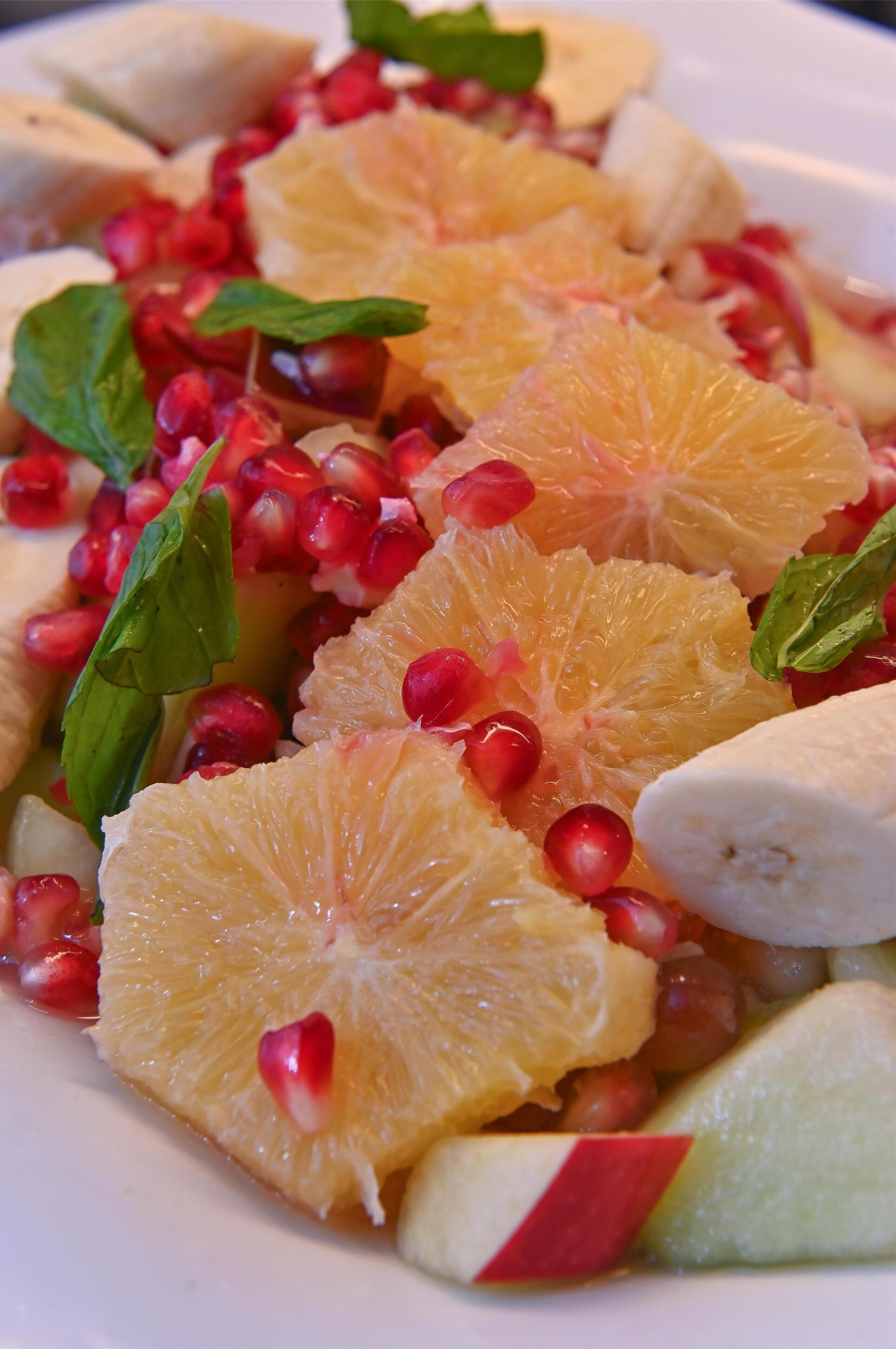 Une salade de fruits contenant des graines de grenade, des tranches d’orange, des morceaux de banane et des feuilles de menthe.