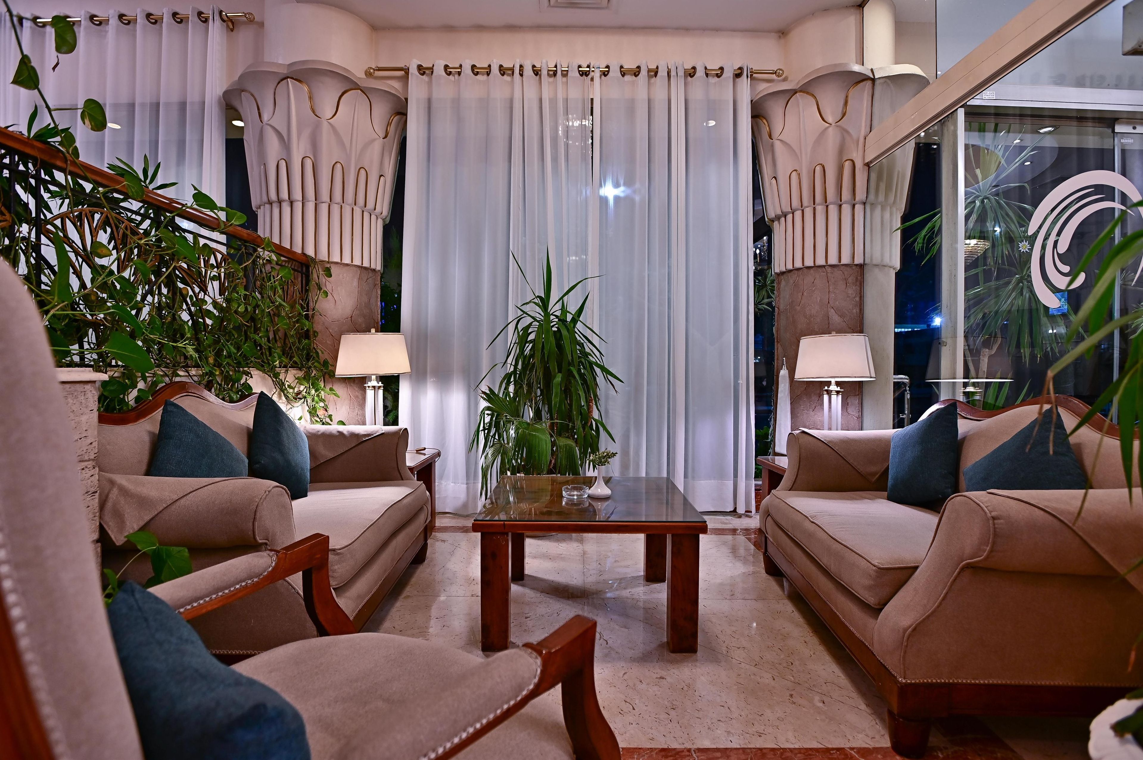 A well-lit, elegantly furnished living room with beige sofas, a glass coffee table, and potted plants.
