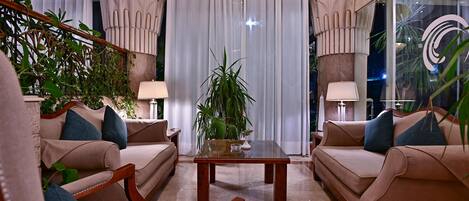 A well-lit, elegantly furnished living room with beige sofas, a glass coffee table, and potted plants.