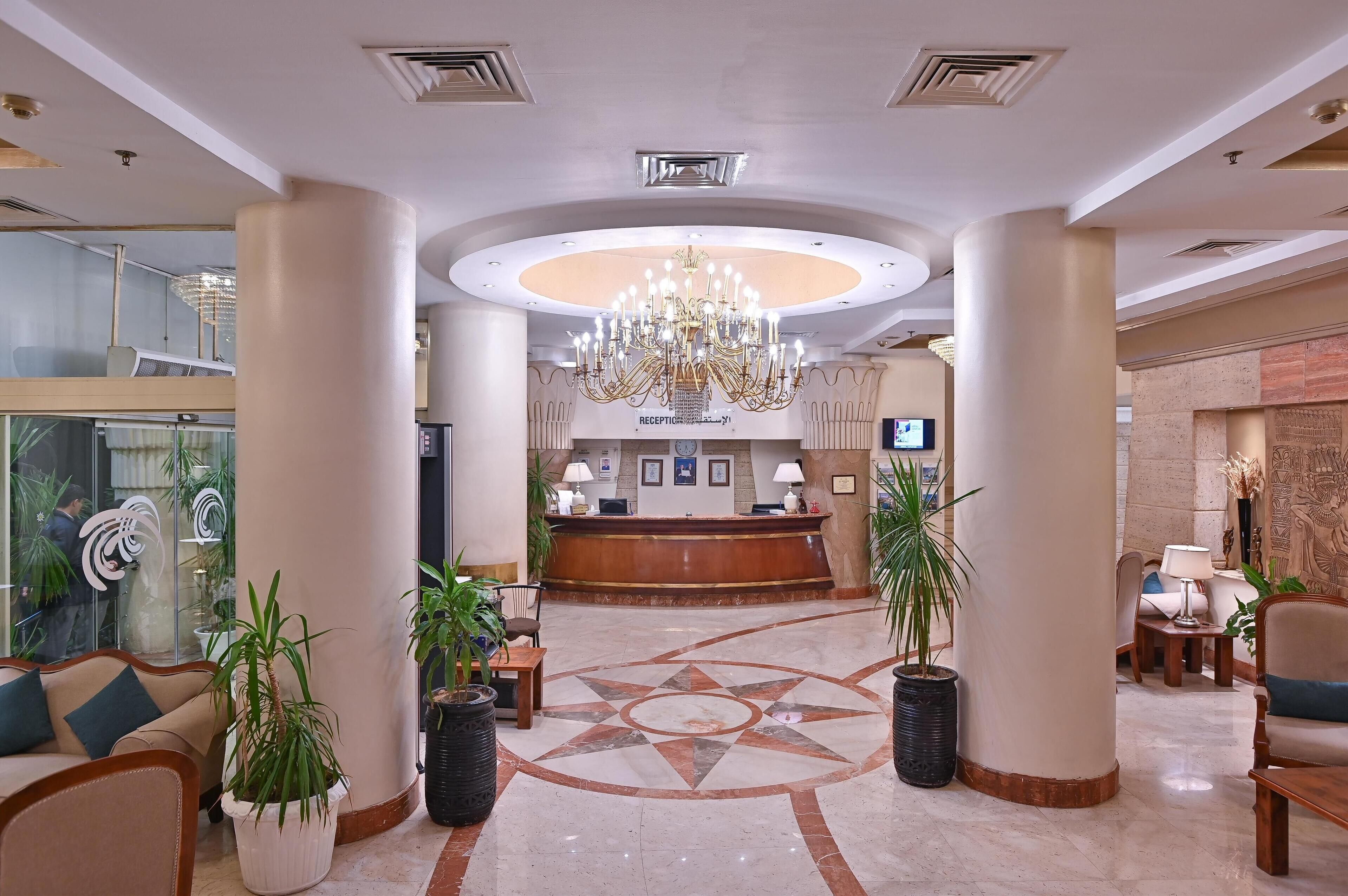 A hotel lobby with a reception desk, a chandelier, potted plants, and a patterned floor.