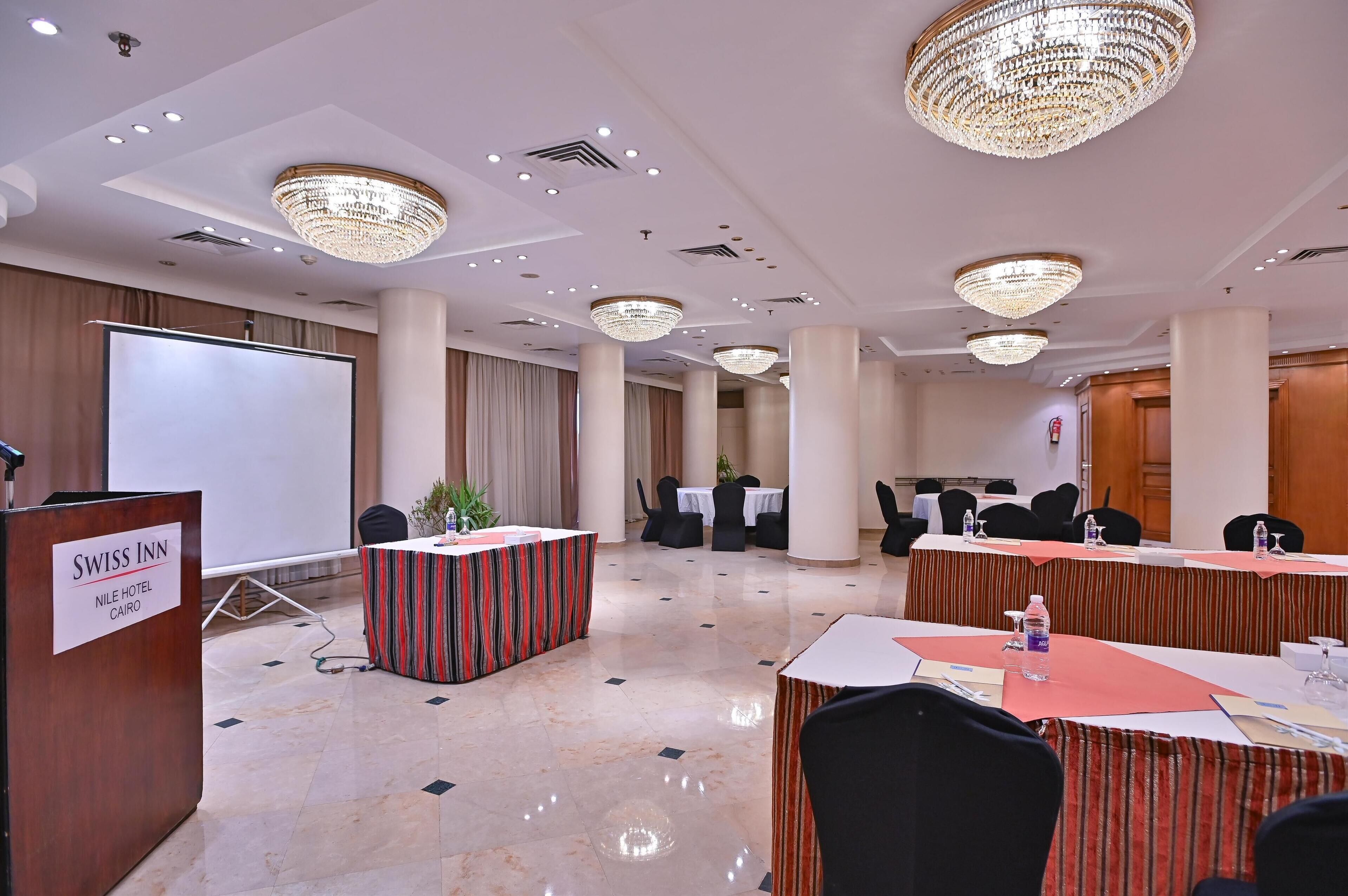 Un salón de conferencias con un atril que dice "SWISS INN", mesas, sillas y una pantalla grande.