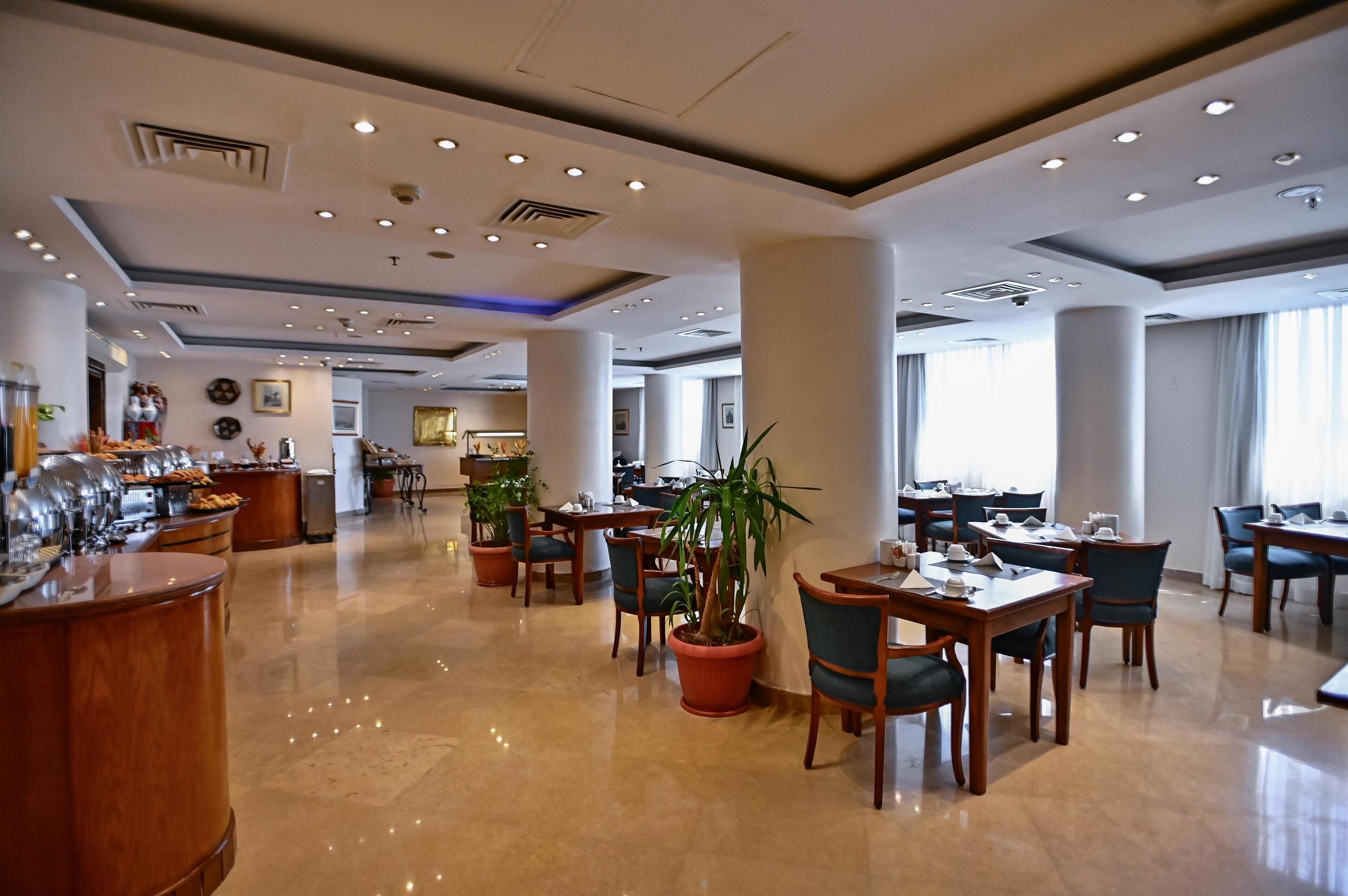 A hotel dining area with multiple tables and chairs, a buffet counter, and potted plants.