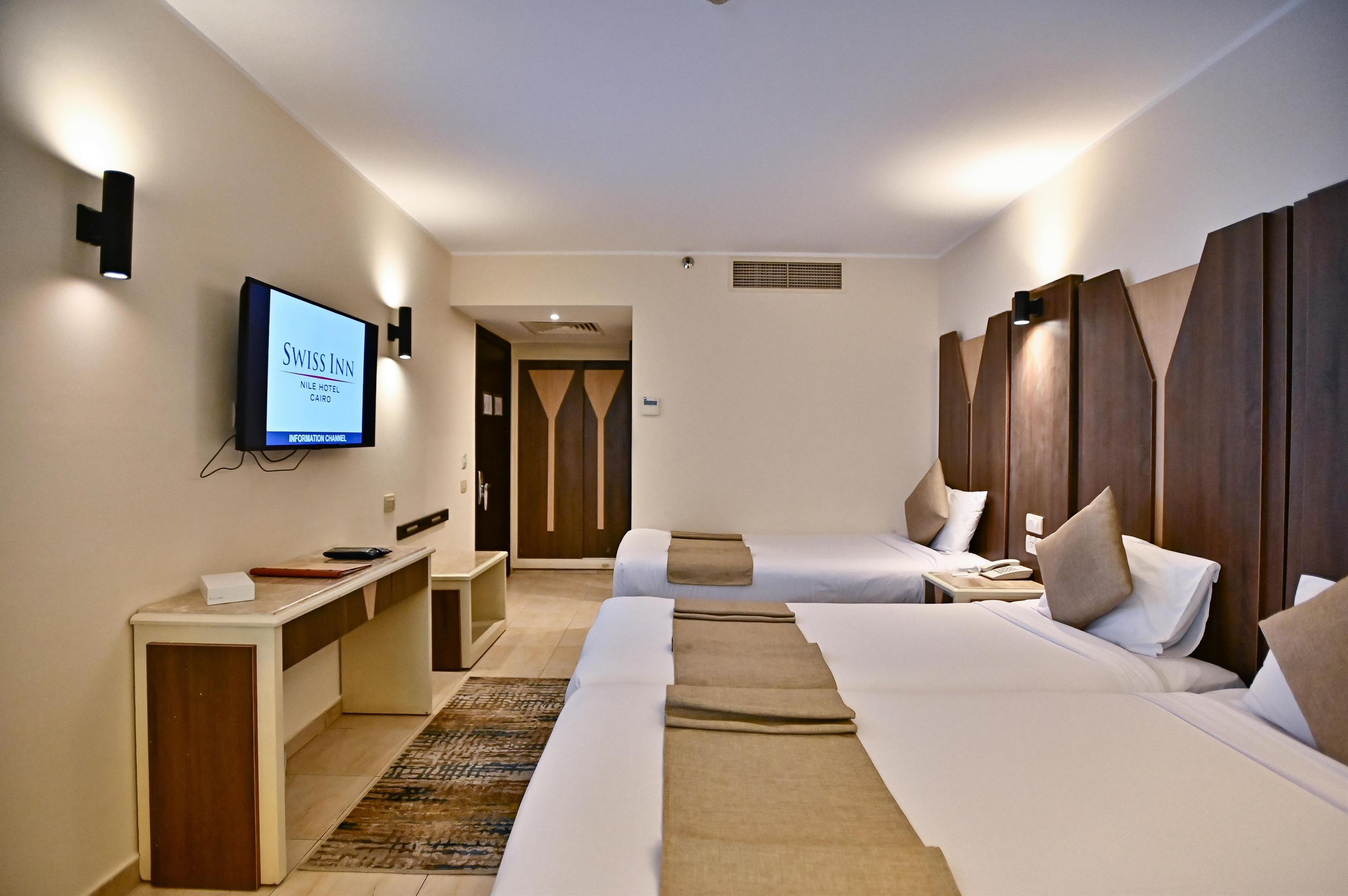 A hotel room with two beds, a desk, and a TV displaying the hotel name.
