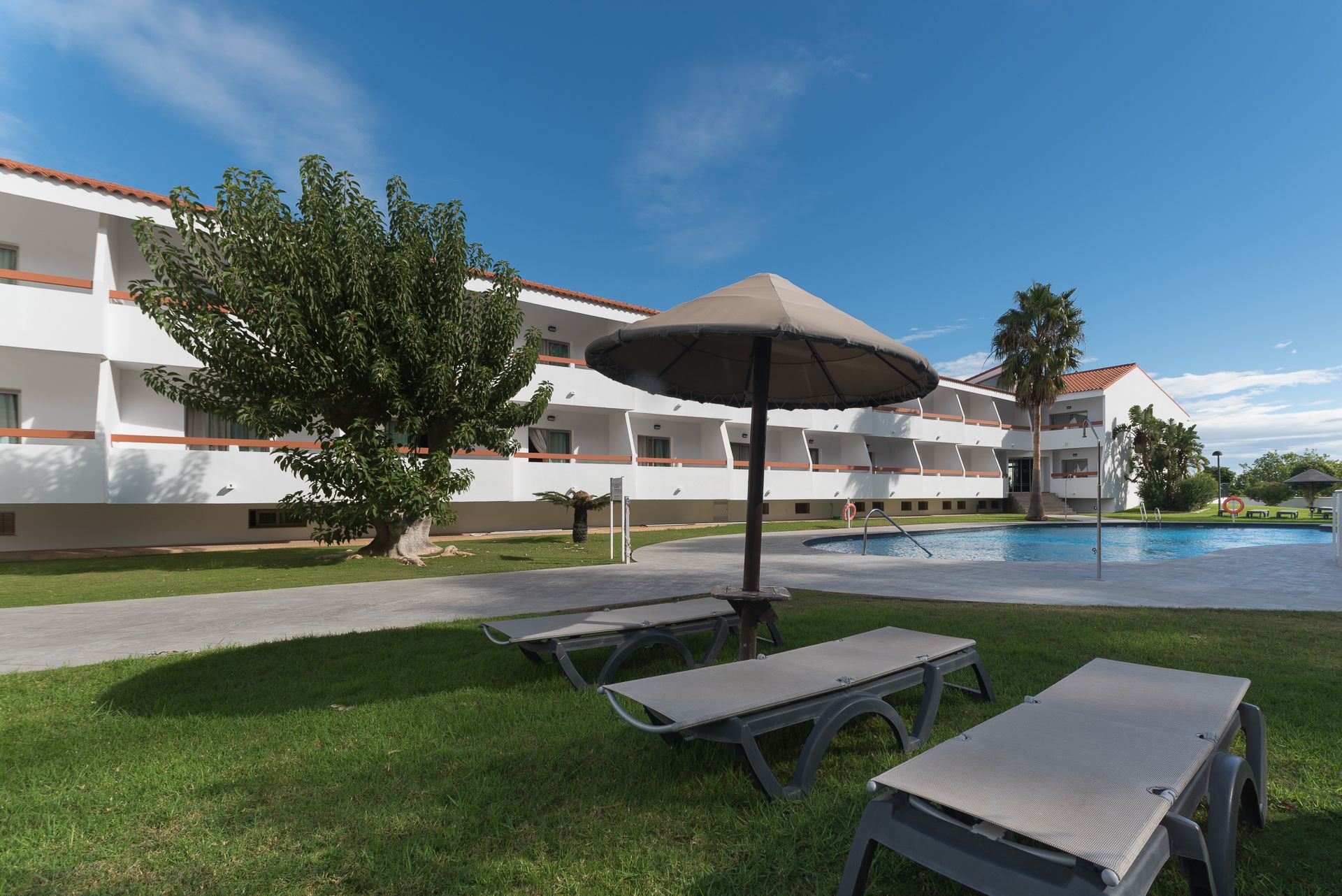 A poolside area with lounge chairs and an umbrella, a building with balconies, and a clear blue sky.