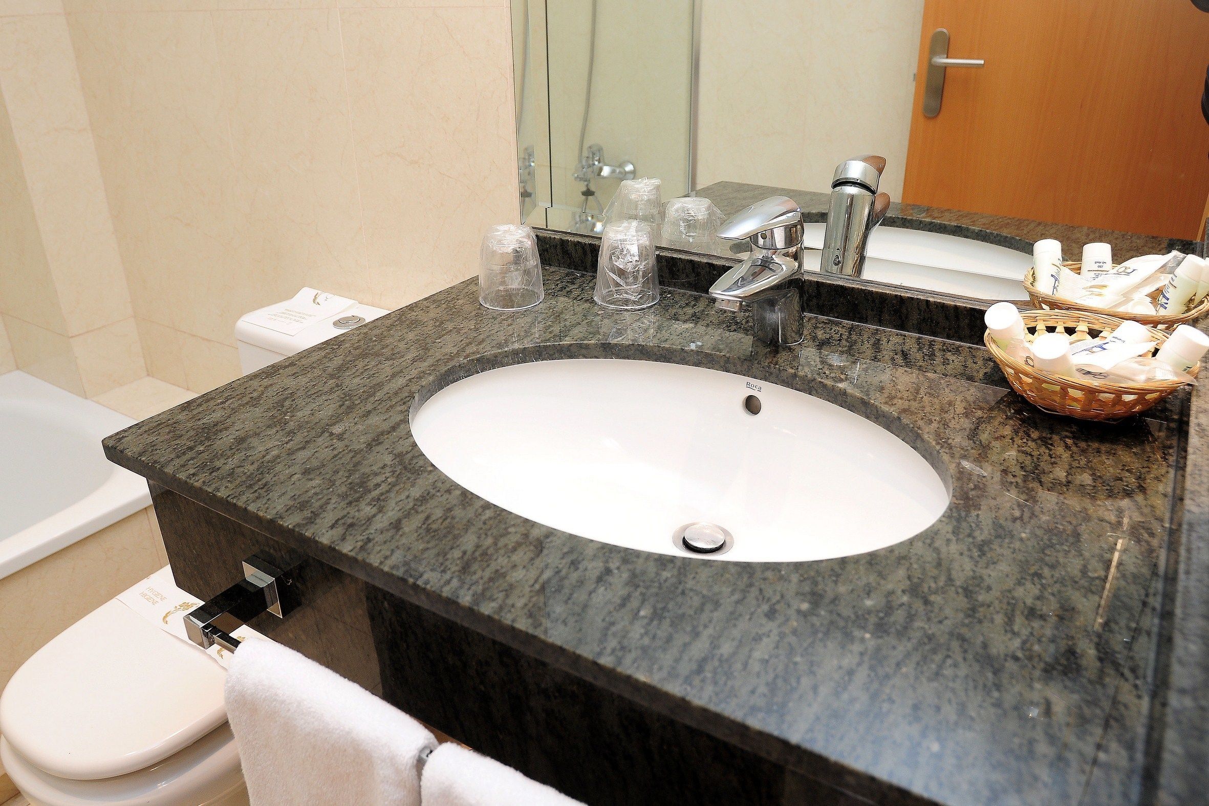 Un baño con encimera de granito, un lavabo blanco, un espejo, un inodoro y una cesta de artículos de aseo.