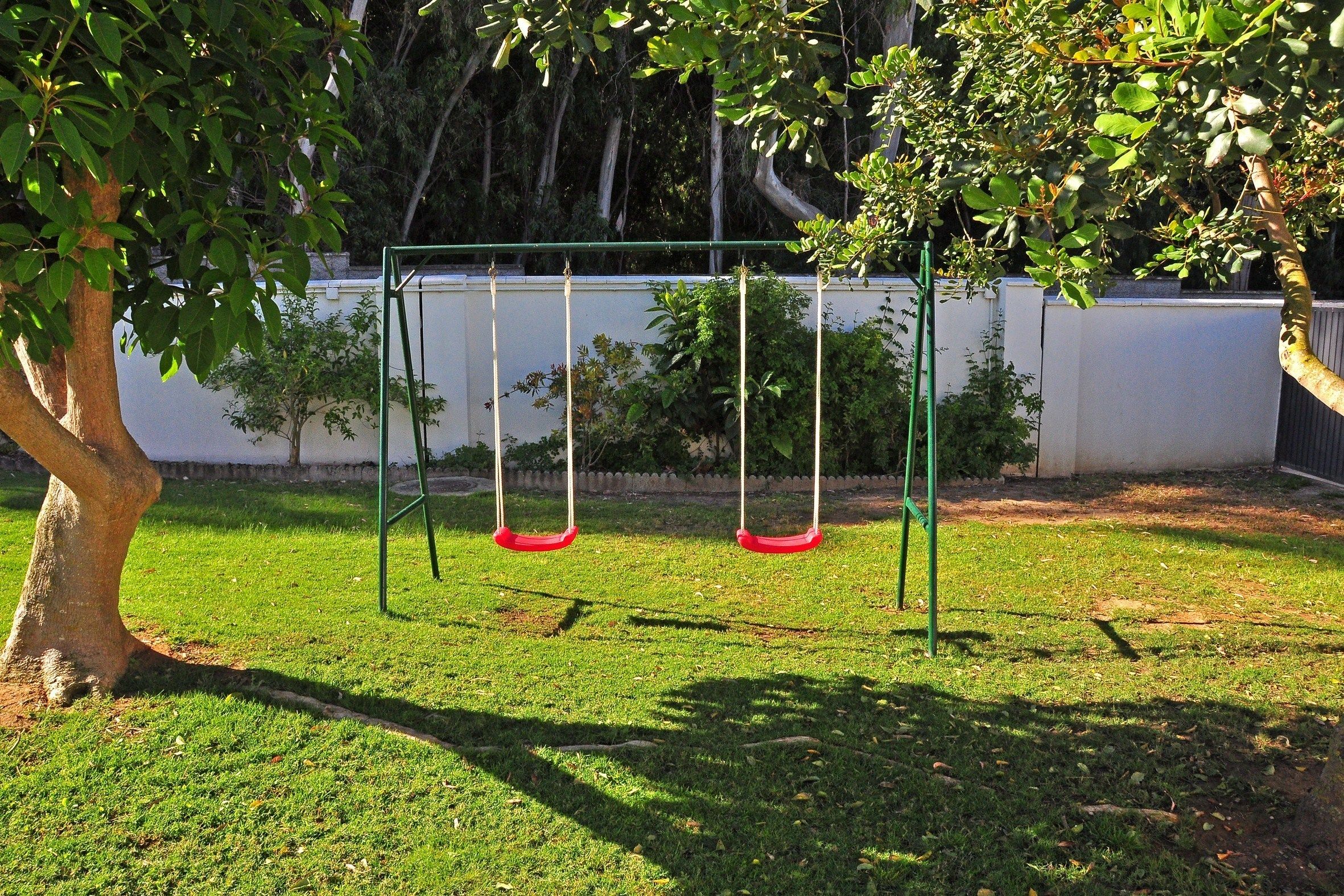 A swing set in a grassy area with a tree and a white wall in the background.