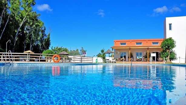 Una piscina con agua cristalina azul, rodeada por una terraza de madera y un edificio blanco con techo rojo.