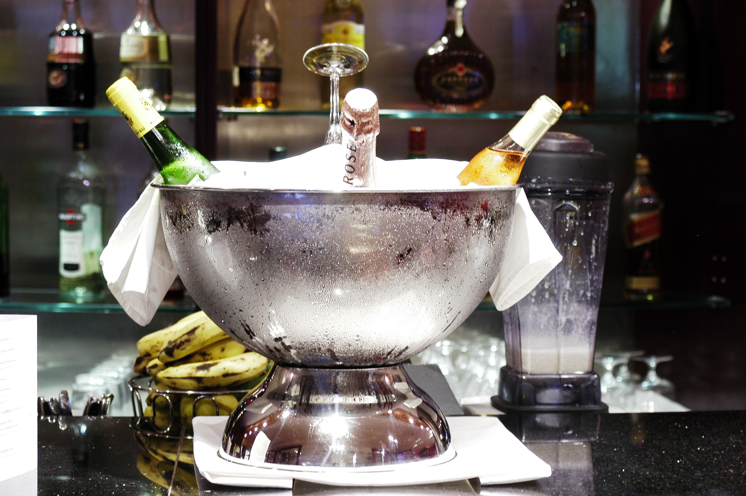 A bar setting with a large metallic bowl containing champagne bottles, a glass, and a banana on a reflective surface.