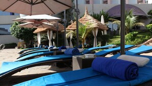 Une rangée de chaises longues dotées de coussins bleus et de serviettes blanches, disposées sous des parasols en chaume, avec une piscine et des palmiers en arrière-plan.