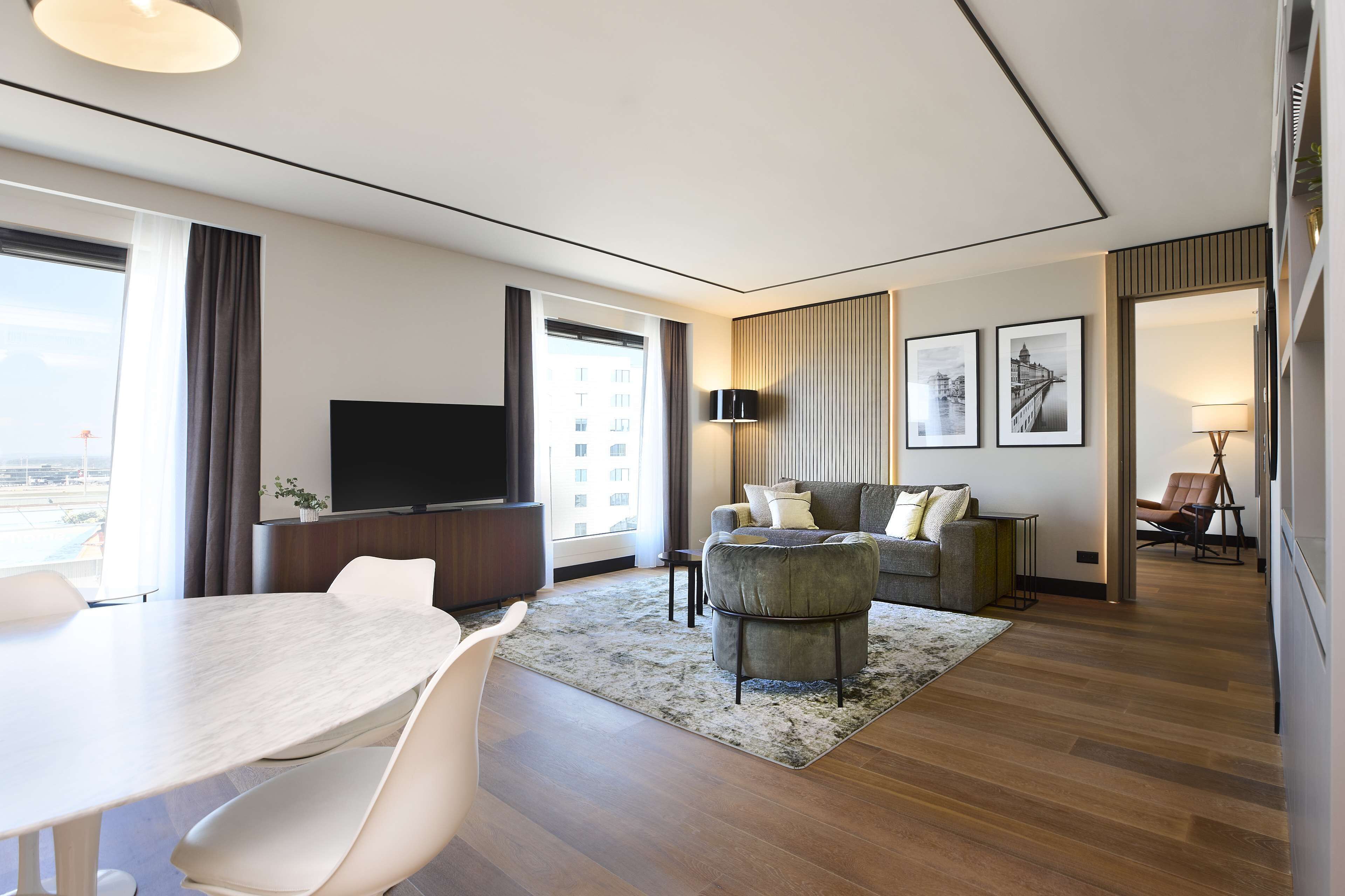 Une chambre d’hôtel moderne comprenant un coin repas, un salon avec un canapé et des fauteuils, une télévision à écran plat et de grandes fenêtres offrant une vue sur la ville.