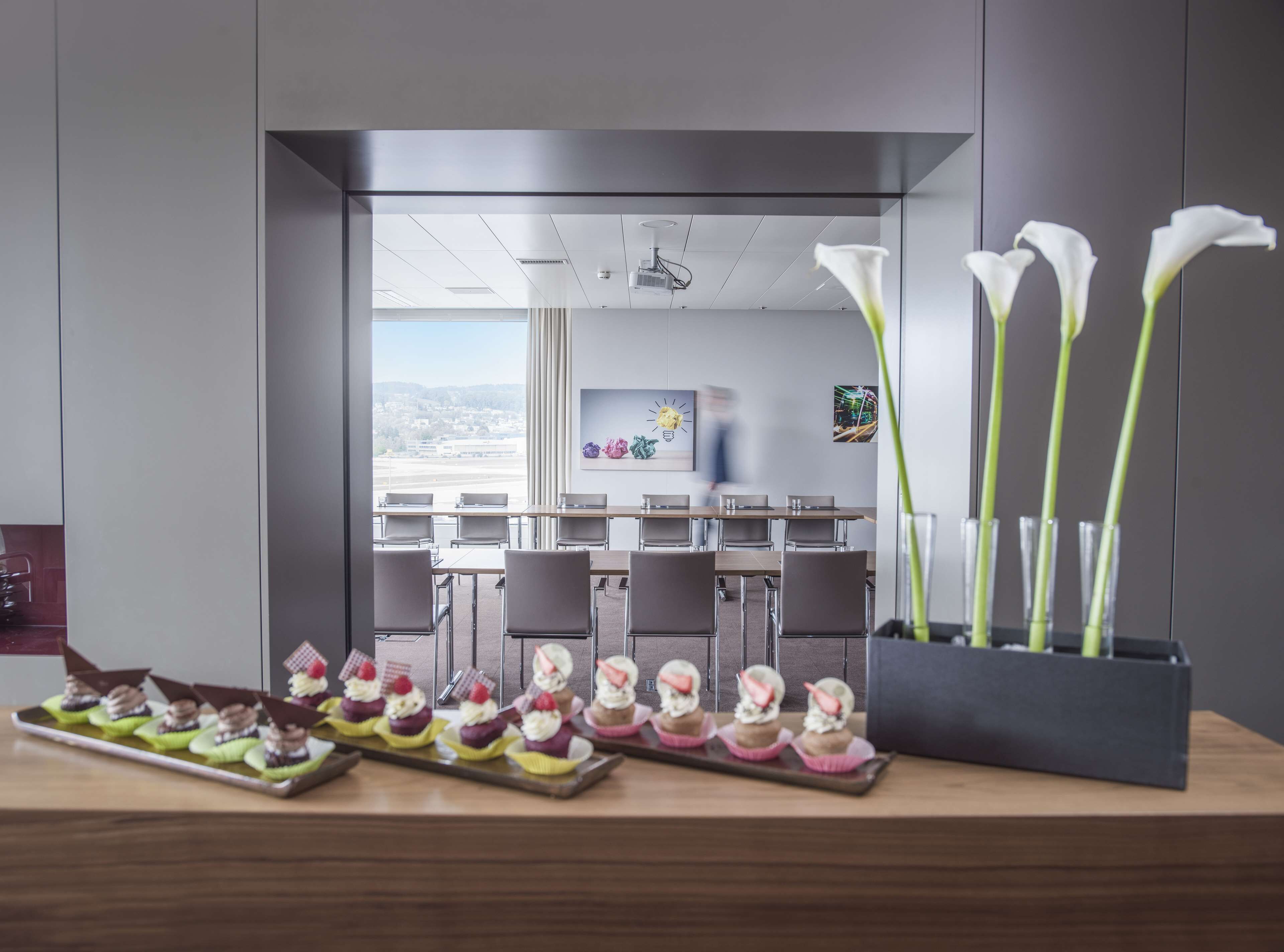 A modern conference room with a view of a cityscape, a glass partition, and a table with cupcakes and calla lilies.