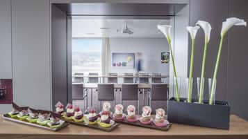 A modern conference room with a view of a cityscape, a glass partition, and a table with cupcakes and calla lilies.