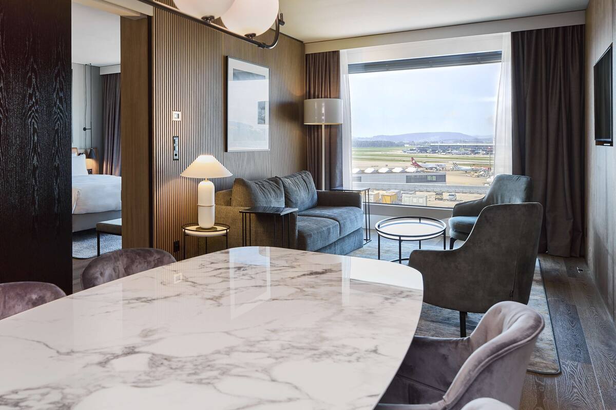Habitación de hotel con una amplia mesa de mármol, un sofá, una pequeña mesa redonda y vistas al aeropuerto.