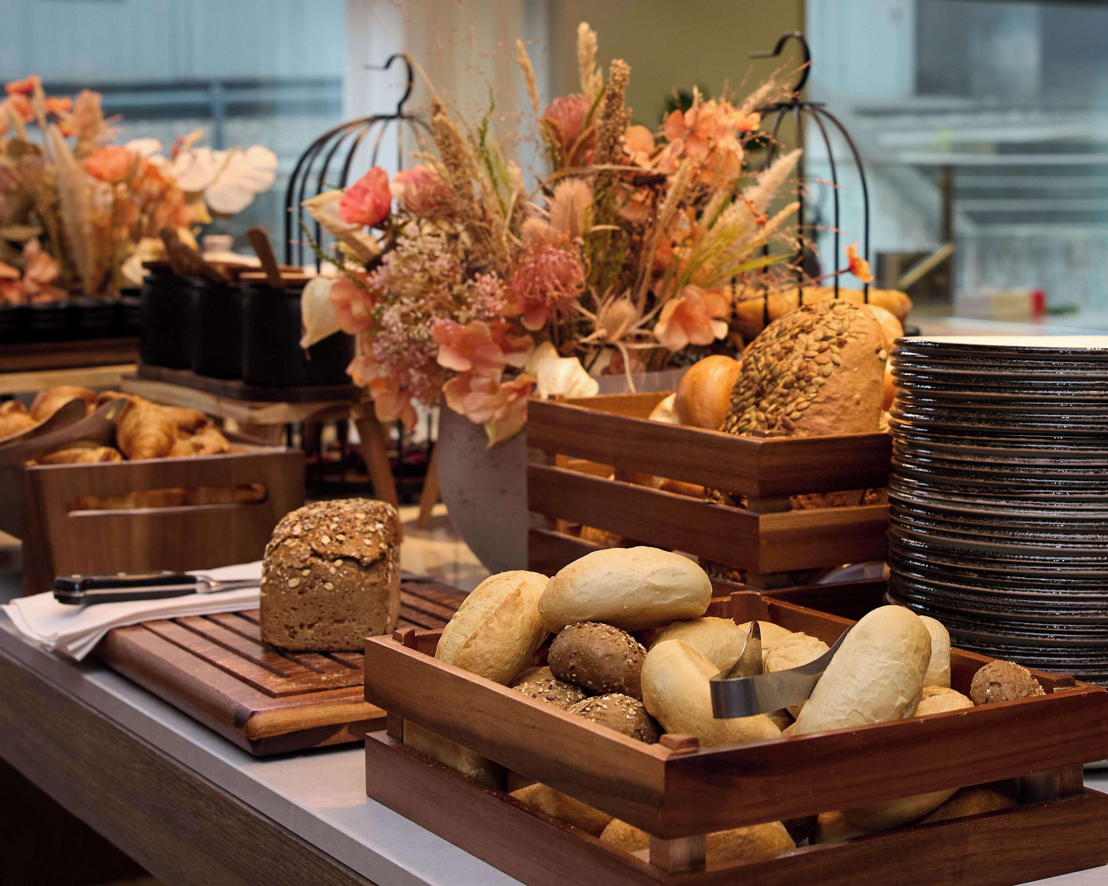 A buffet table with various breads and pastries in wooden trays, accompanied by floral centerpieces.