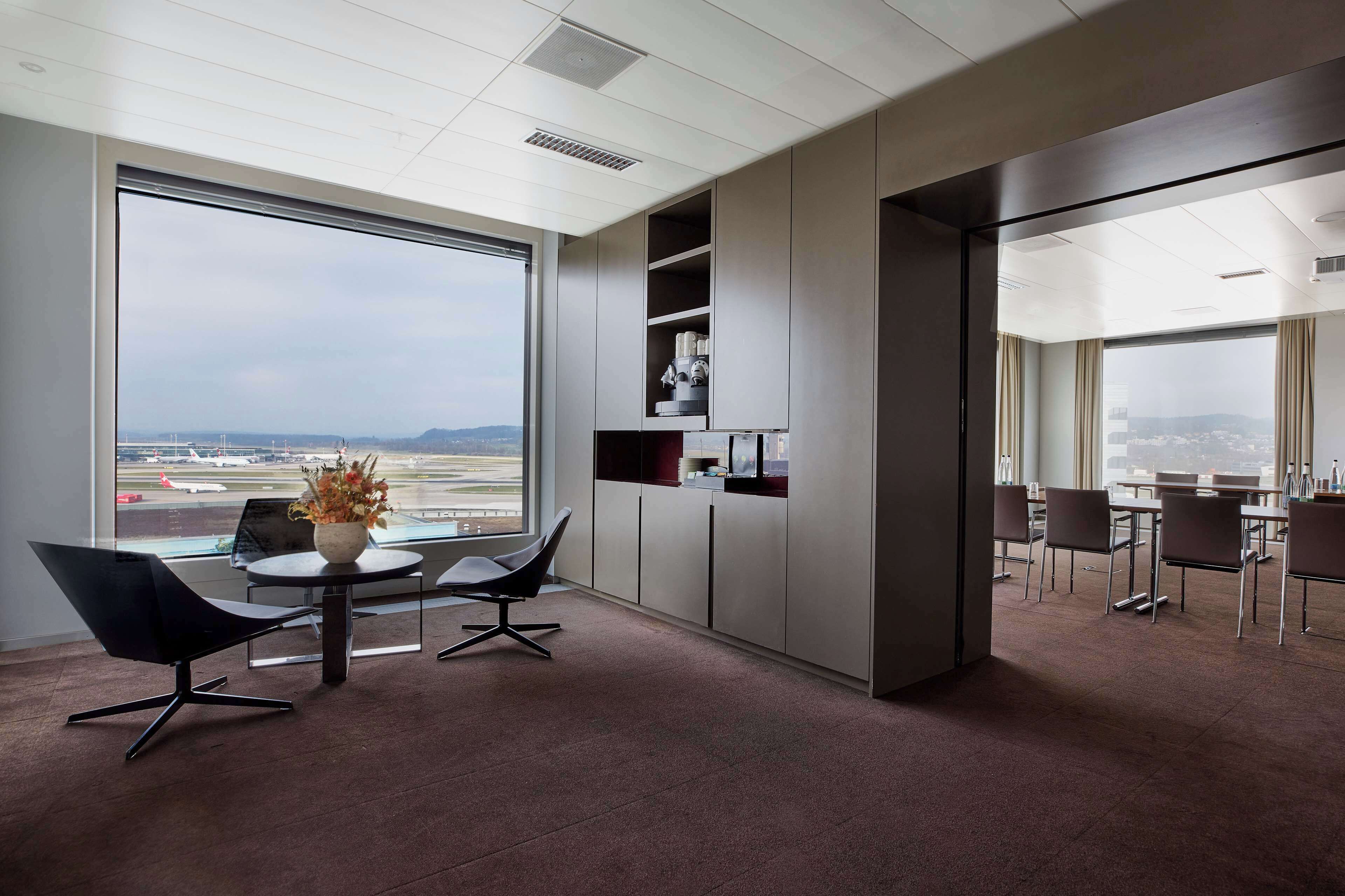Ein moderner Arbeitsbereich mit einem großen Fenster, das Blick auf einen Flughafen und die Stadt bietet. Zwei schwarze Stühle und ein kleiner Tisch mit einem Blumenstrauß sind vorhanden.