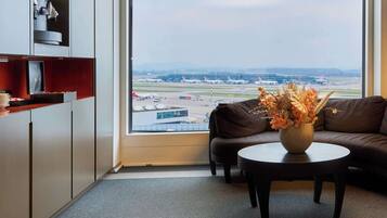 A modern room with a large window offering a view of an airport. There is a brown leather sofa, a small round table with a vase of dried flowers, and a cabinet with a coffee machine.