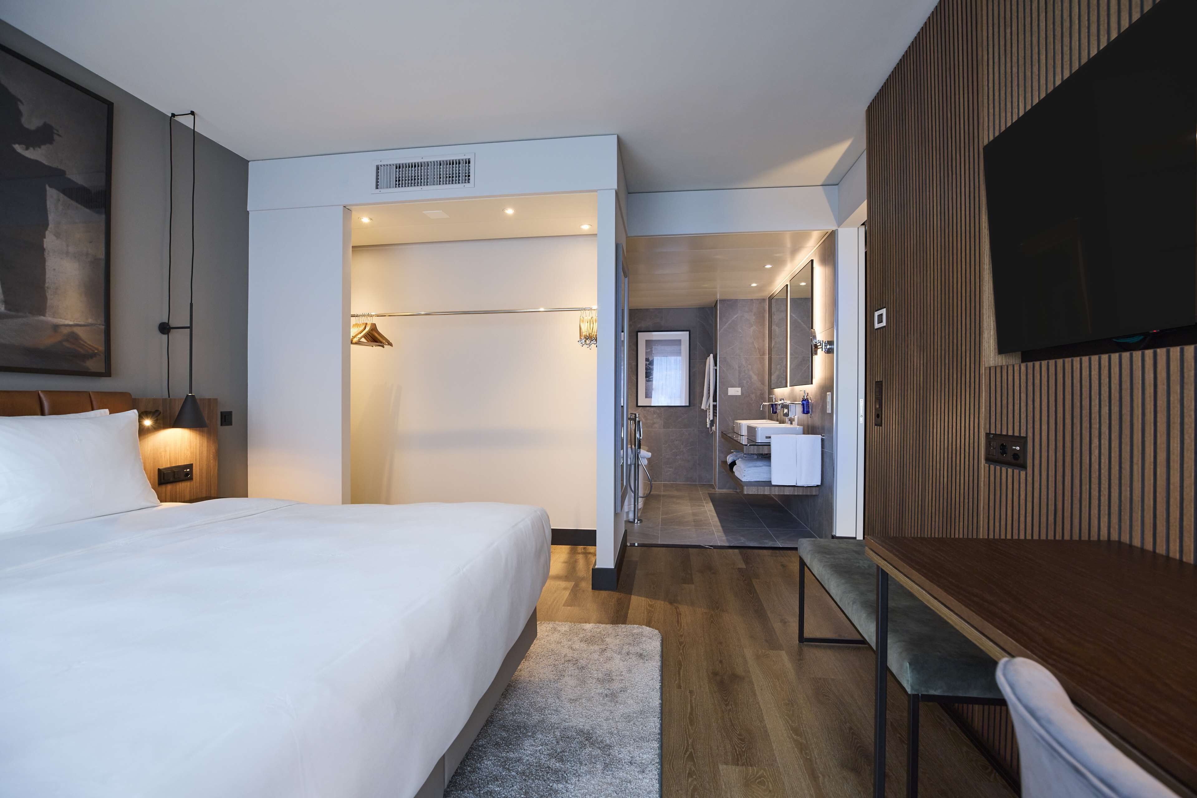 A modern hotel room with a large bed, a desk, a TV, and a bathroom visible through an open door.
