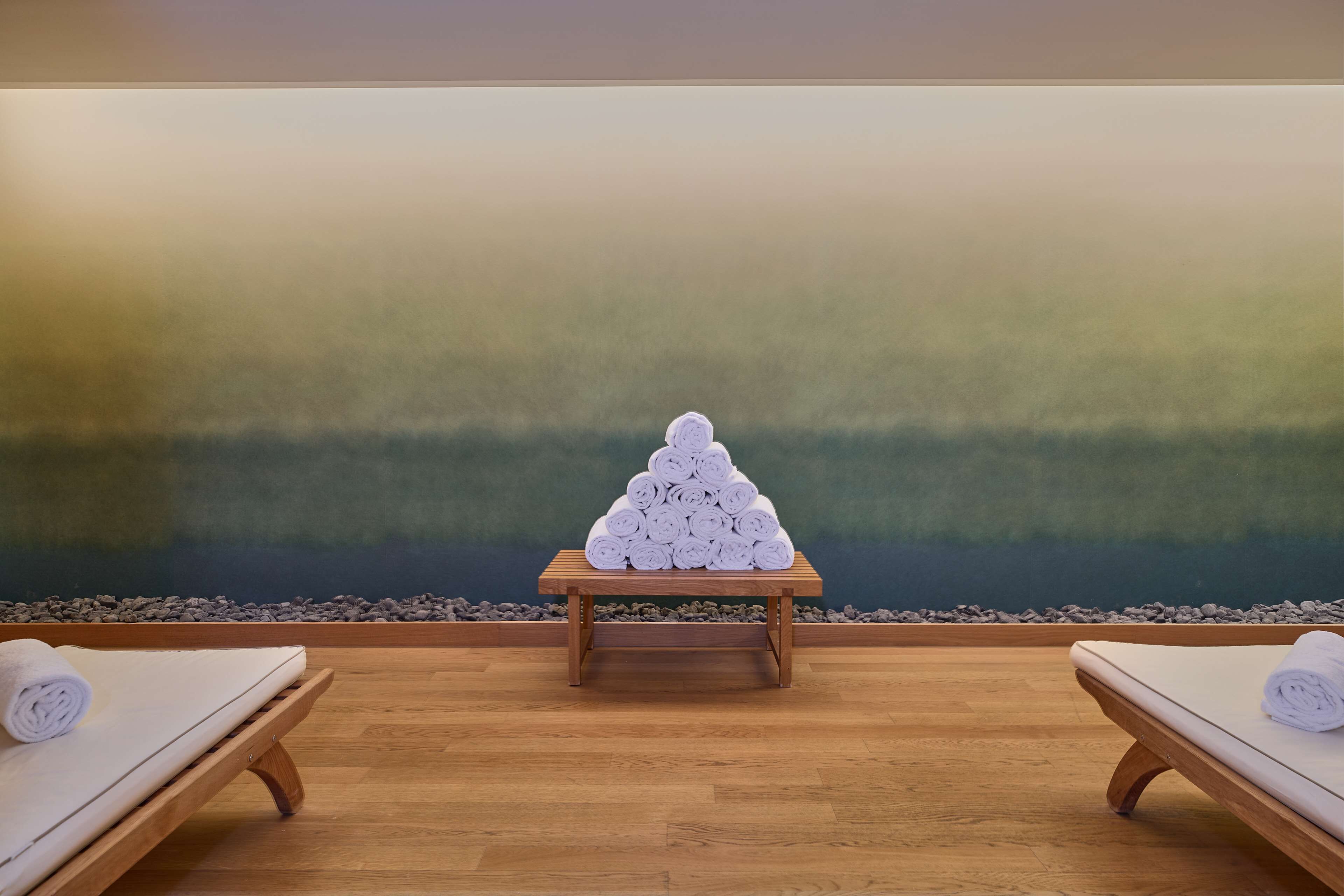 A spa room with two wooden massage tables, a wooden table with rolled towels in the center, and a large abstract wall mural.