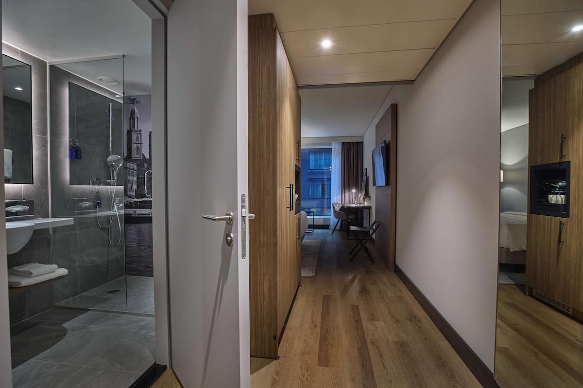 Una habitación de hotel moderna con ducha de vidrio, piso de madera, zona de comedor con mesa y sillas, y televisor montado en la pared.