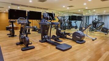 A gym with various exercise machines, a wooden floor, and a large mirror reflecting the equipment.