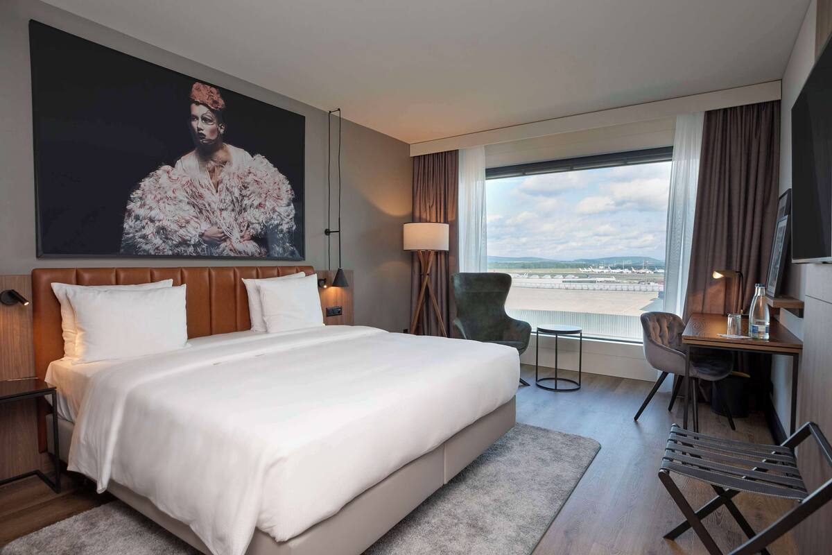 Habitación de hotel con una cama grande, un escritorio, una silla, un televisor y un ventanal con vistas.