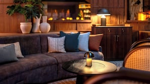 Un área de estar moderna con un sofá de color oscuro, una mesa redonda con lámpara y una barra de bar al fondo.