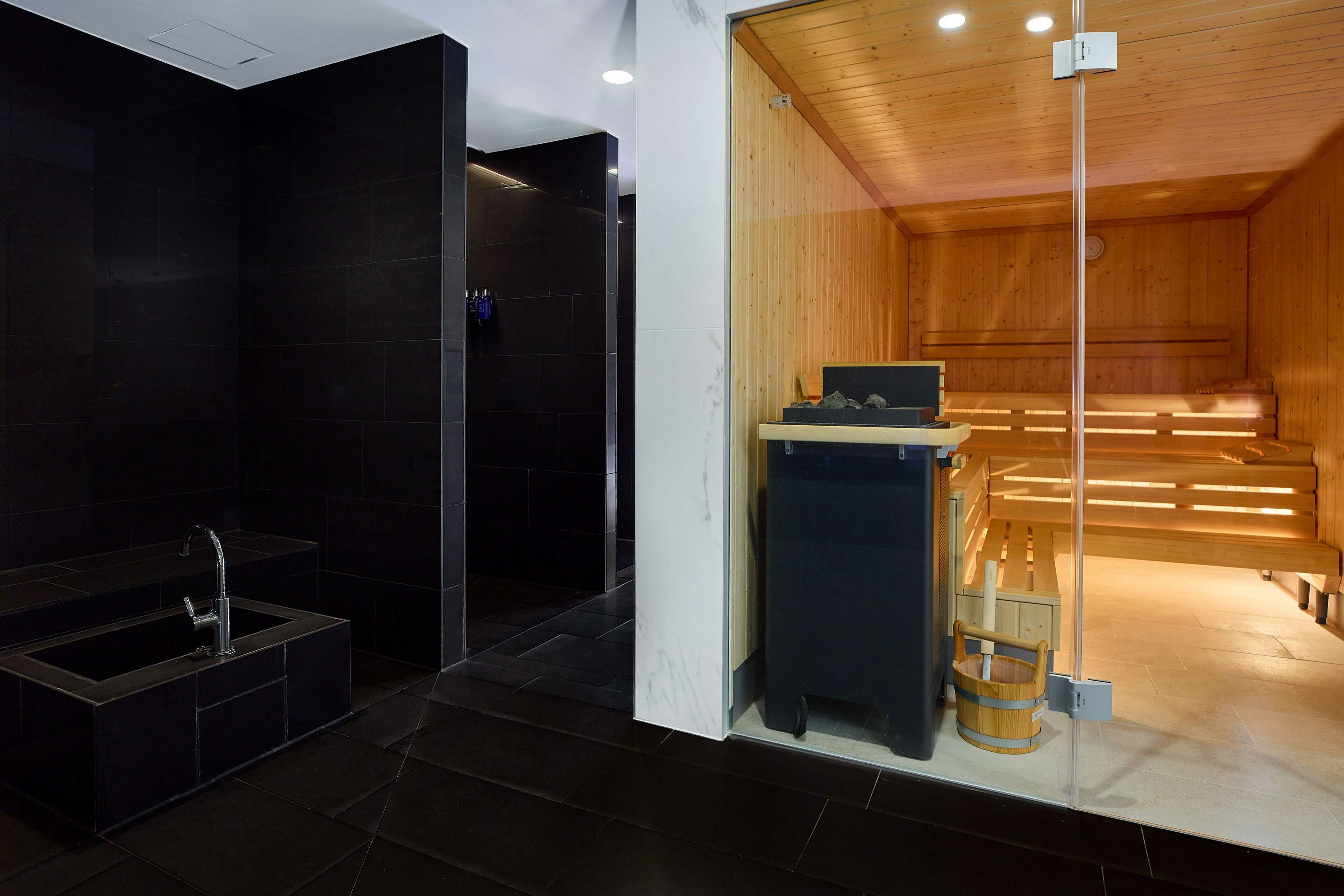 A modern bathroom with a black-tiled shower area and a wooden sauna visible through a glass partition.