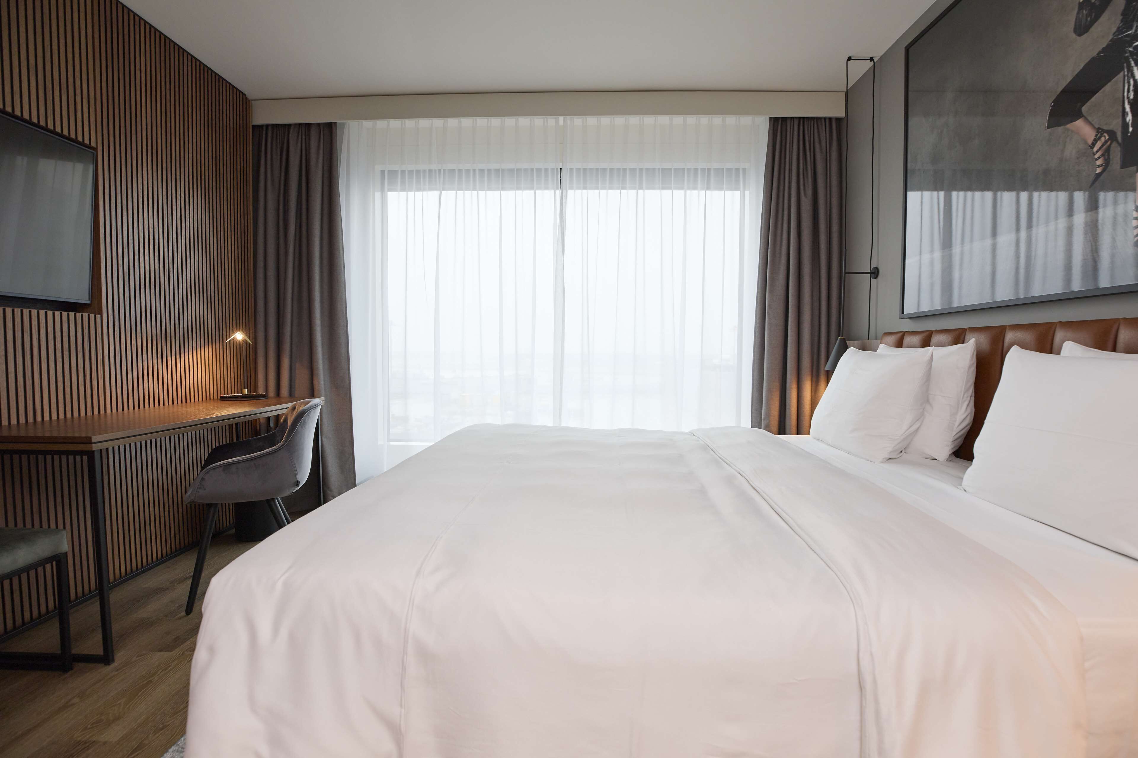 Habitación de hotel con una cama grande, un escritorio con silla, un televisor montado en la pared y un ventanal con cortinas.