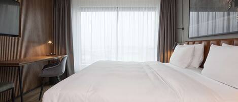 Habitación de hotel con una cama grande, un escritorio con silla, un televisor montado en la pared y un ventanal con cortinas.