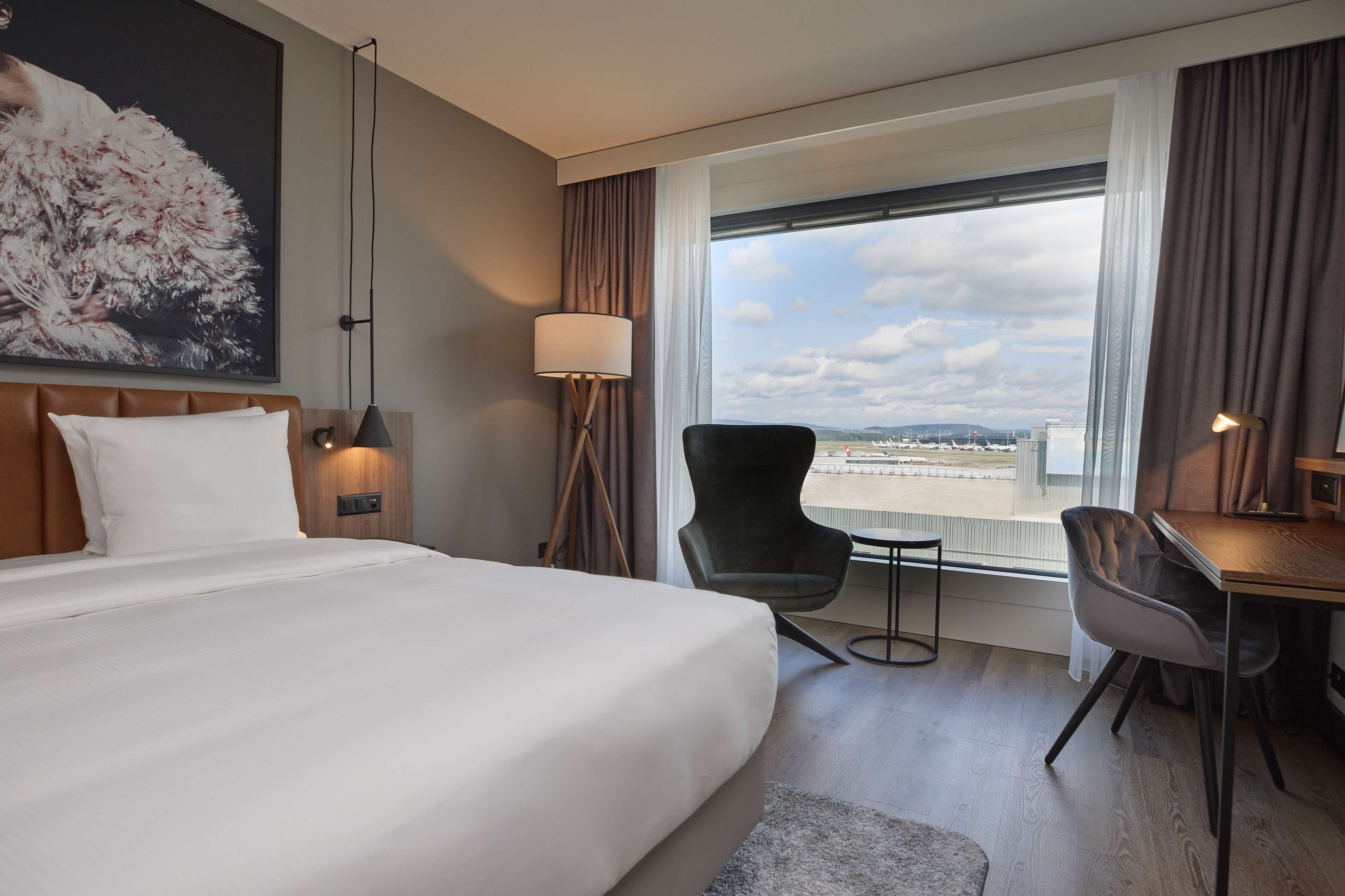 A hotel room with a large bed, a desk, a chair, a lamp, and a view of the city through the window.