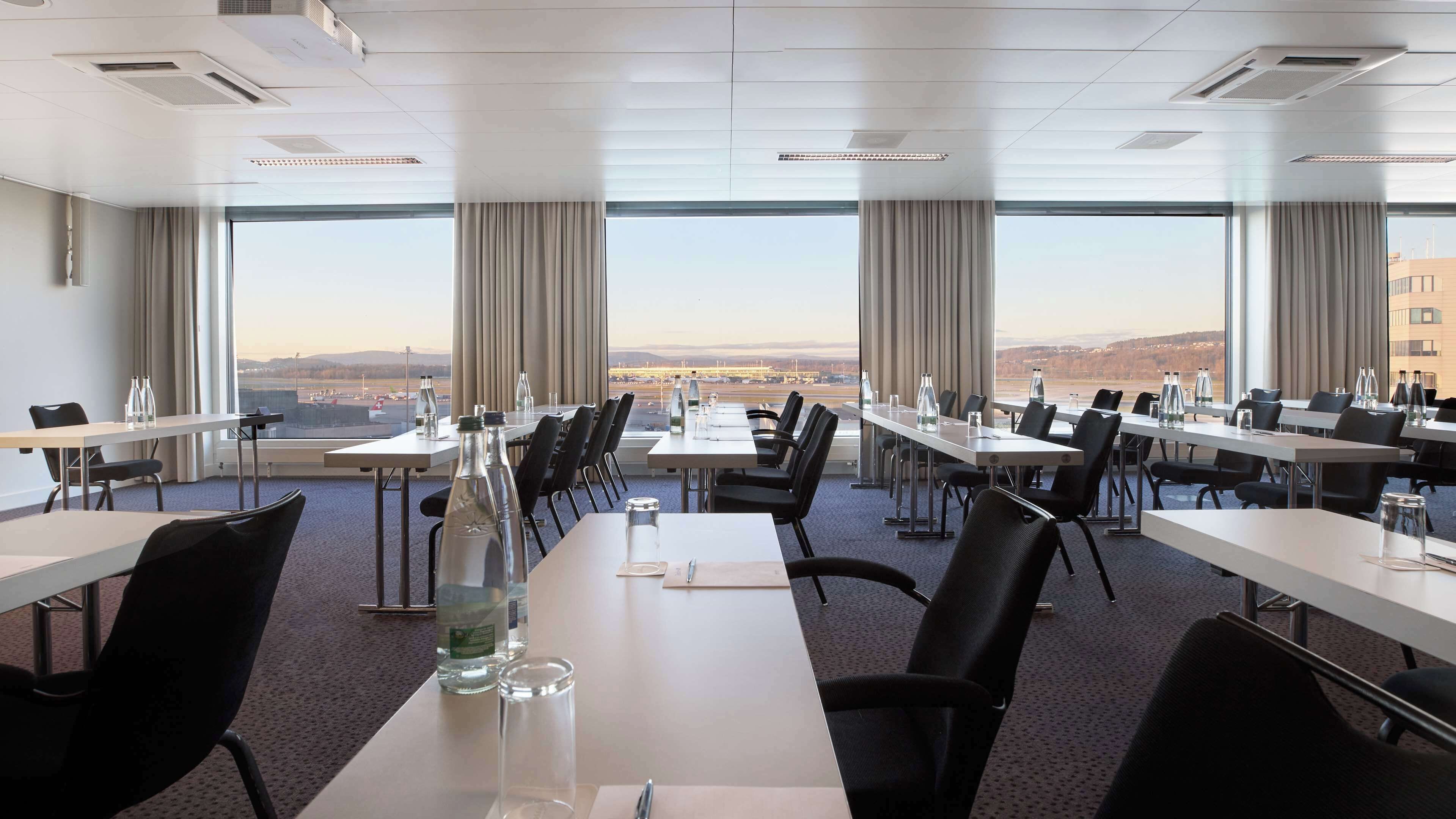 Una sala de conferencias con varias mesas y sillas, amplias ventanas que ofrecen vistas a un paisaje urbano, y un diseño de interiores moderno.