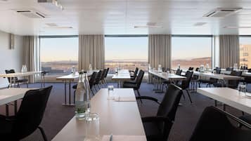 Una sala de conferencias con varias mesas y sillas, amplias ventanas que ofrecen vistas a un paisaje urbano, y un diseño de interiores moderno.