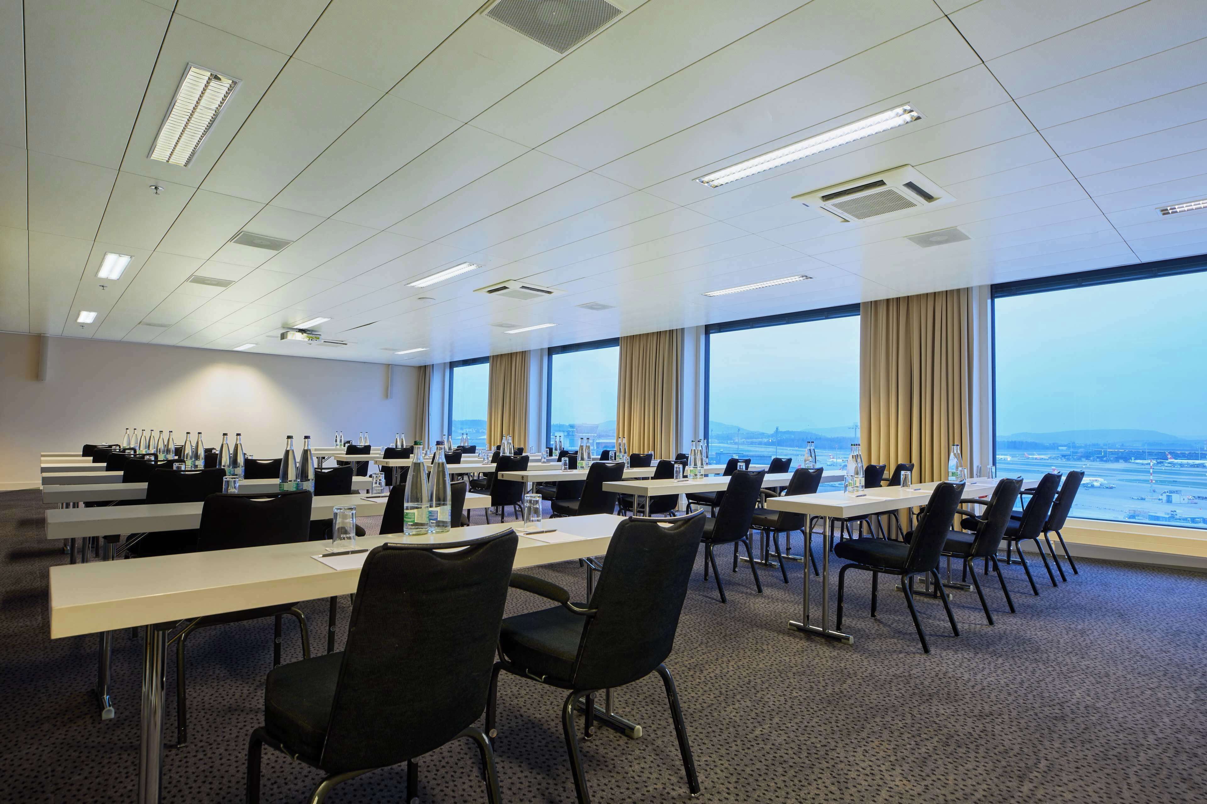 A conference room with multiple tables and chairs, large windows with a view of the cityscape, and a carpeted floor.