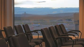 A row of dark-colored chairs facing a large window with a view of an airport and distant mountains.
